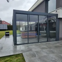 Nowoczesna pergola z szarymi ramami i szklanymi ścianami, z jacuzzi w środku. Na zewnątrz szara kostka brukowa, trawnik i panel słoneczny. Widok na dom.