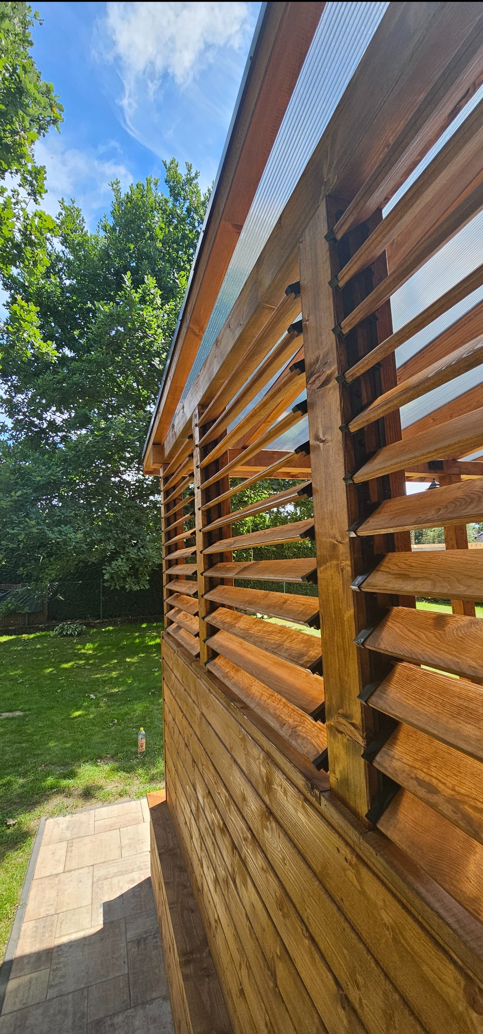 Drewniana konstrukcja z żaluzjami, widok z dołu na tle zieleni ogrodu i błękitnego nieba.