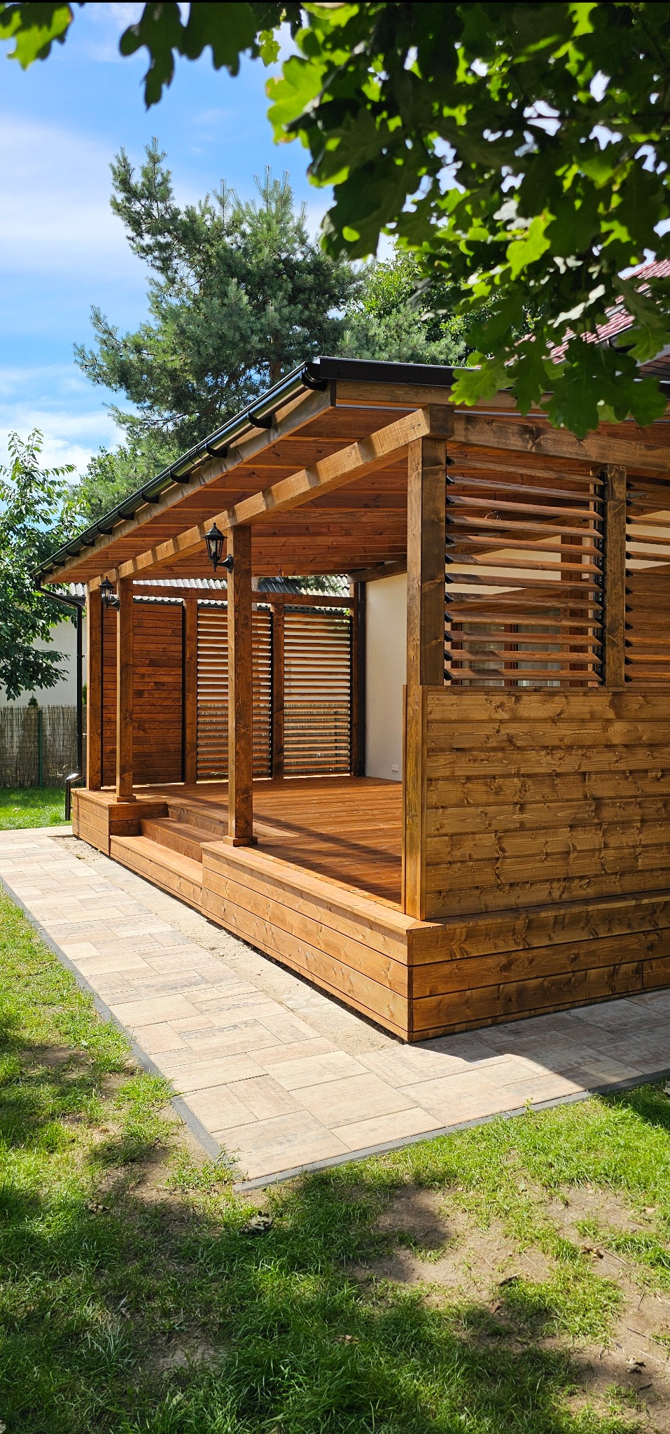 Drewniana pergola z lamelami i oświetleniem zewnętrznym, wykonana w Kobylnicy, z widocznym tarasem i ścieżką z kostki brukowej.