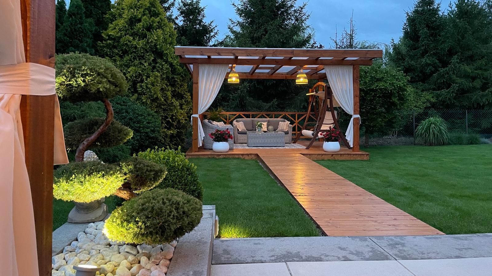 Oświetlona drewniana pergola z huśtawką i zasłonami w ogrodzie, ścieżka z desek prowadzi do platformy z meblami ogrodowymi, dekoracyjne drzewka bonsai.