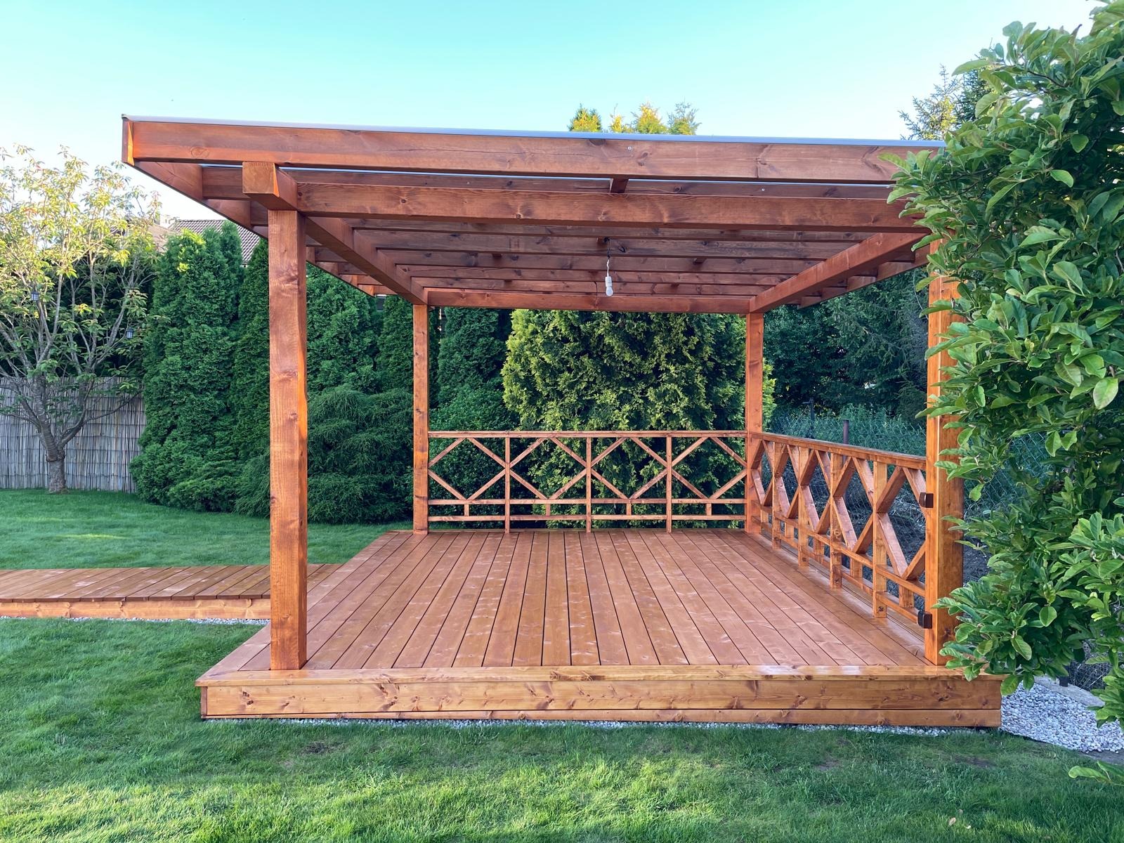 Nowoczesna, zadaszona konstrukcja z drewna z balustradą o dekoracyjnym wzorze rombów, połączona drewnianym chodnikiem z trawnikiem w ogrodzie z iglakami w tle.