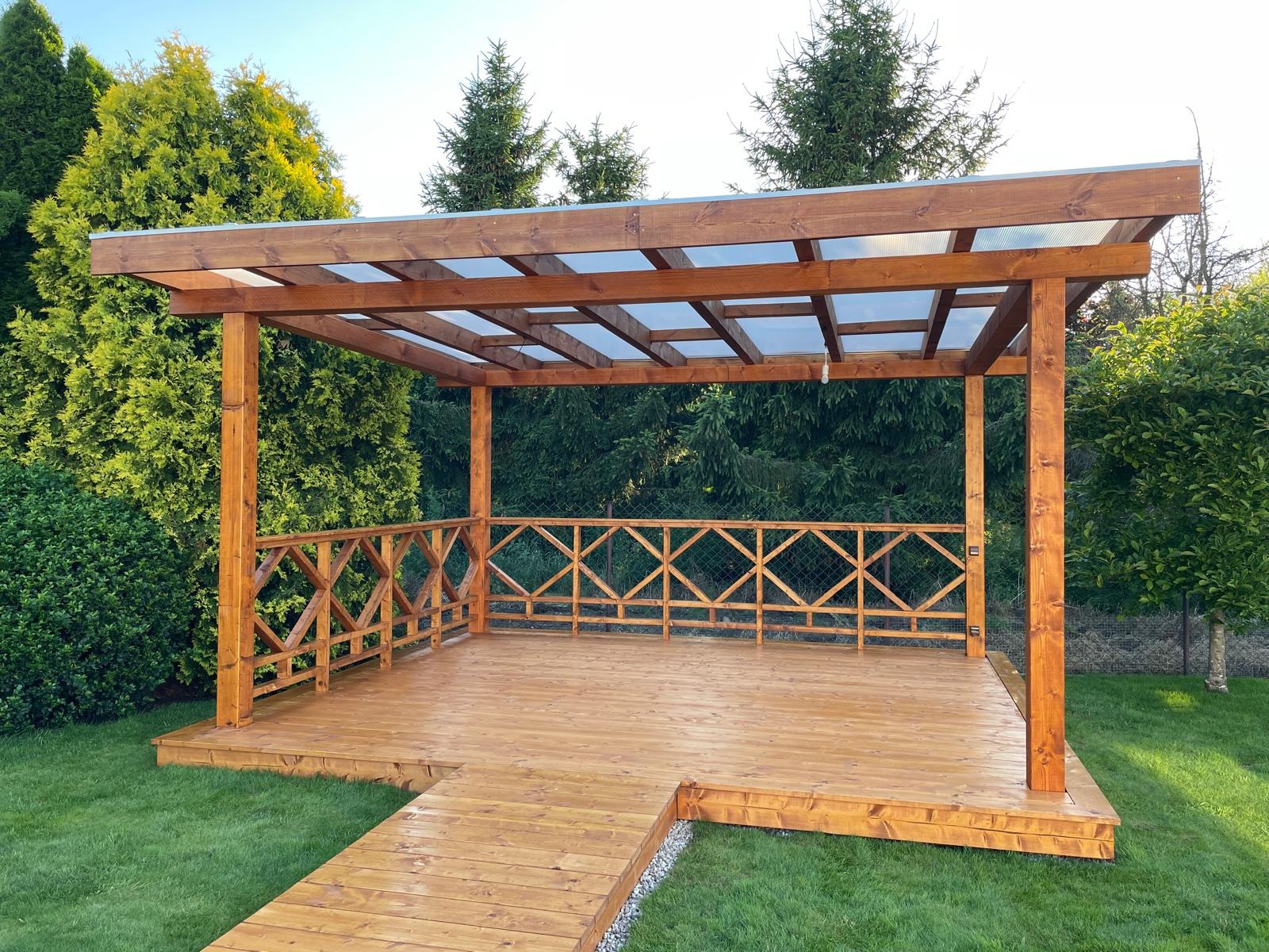 Nowoczesna drewniana konstrukcja z przezroczystym zadaszeniem, z ozdobnymi balustradami i drewnianym podejściem, otoczona zielenią ogrodu.
