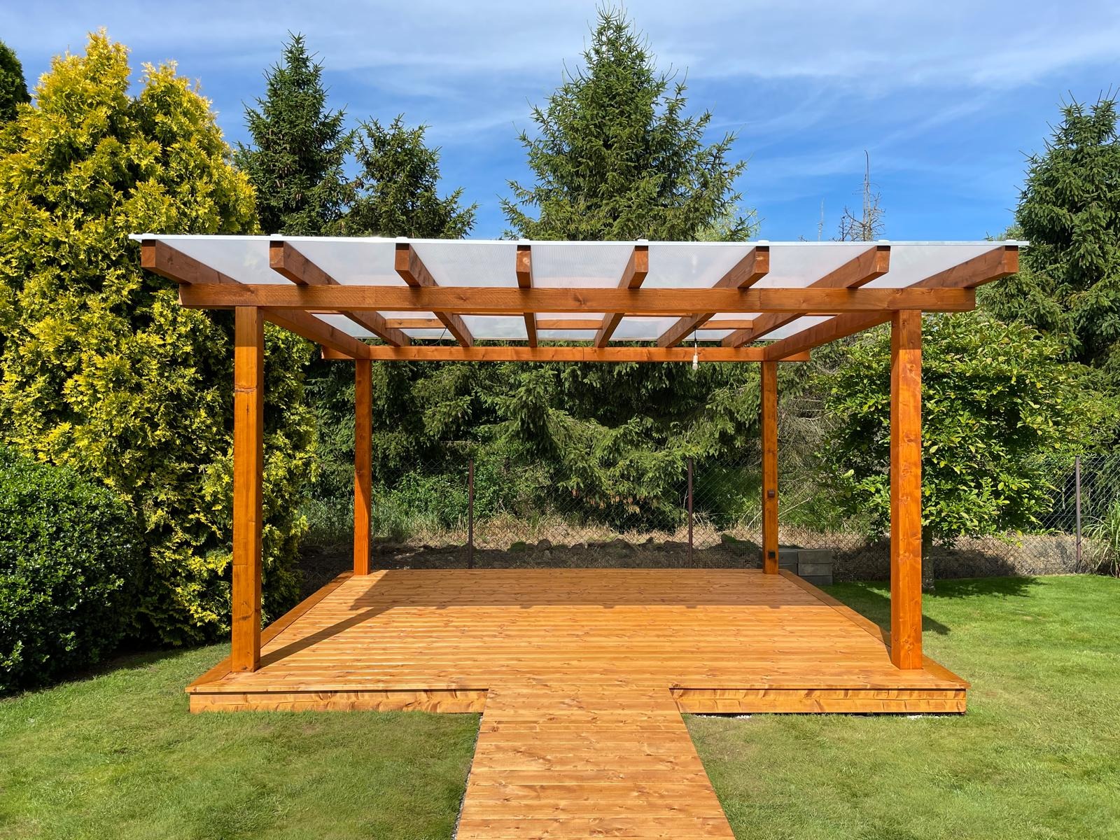 Nowoczesna pergola z drewnianą konstrukcją i przezroczystym zadaszeniem, połączona z tarasem z desek, otoczona zielonym trawnikiem i drzewami w tle.