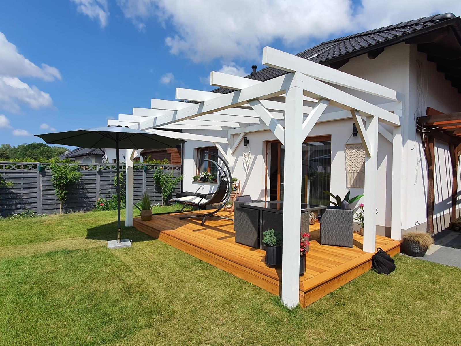 Biała pergola z drewnianym tarasem, na którym stoją meble ogrodowe i huśtawka. Widok na zadbany trawnik i ogrodzenie w słoneczny dzień.