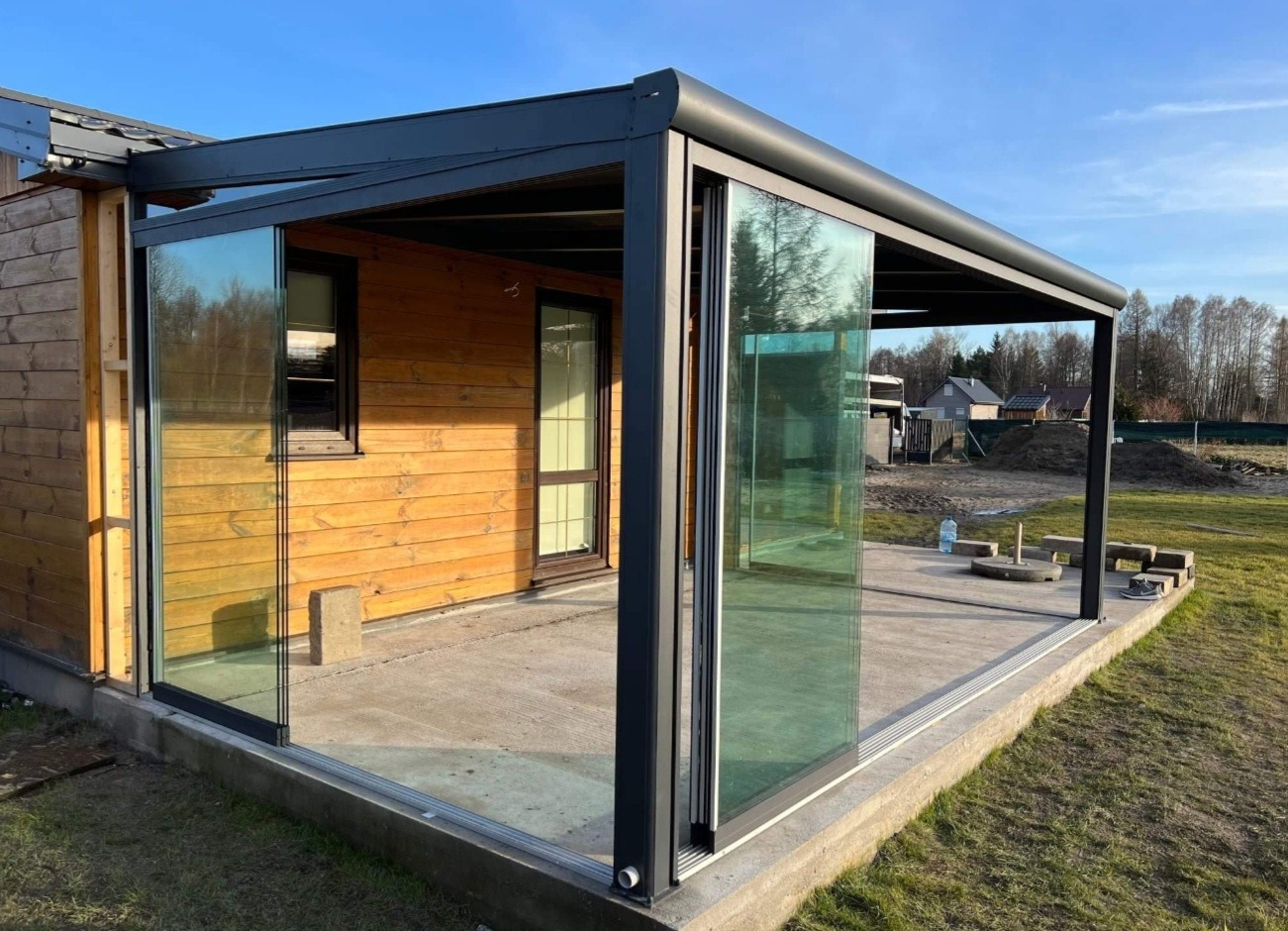 Nowoczesna, czarna pergola metalowa z przesuwnymi szklanymi ścianami, przylegająca do drewnianego domu. Widok na betonowy taras i otoczenie.