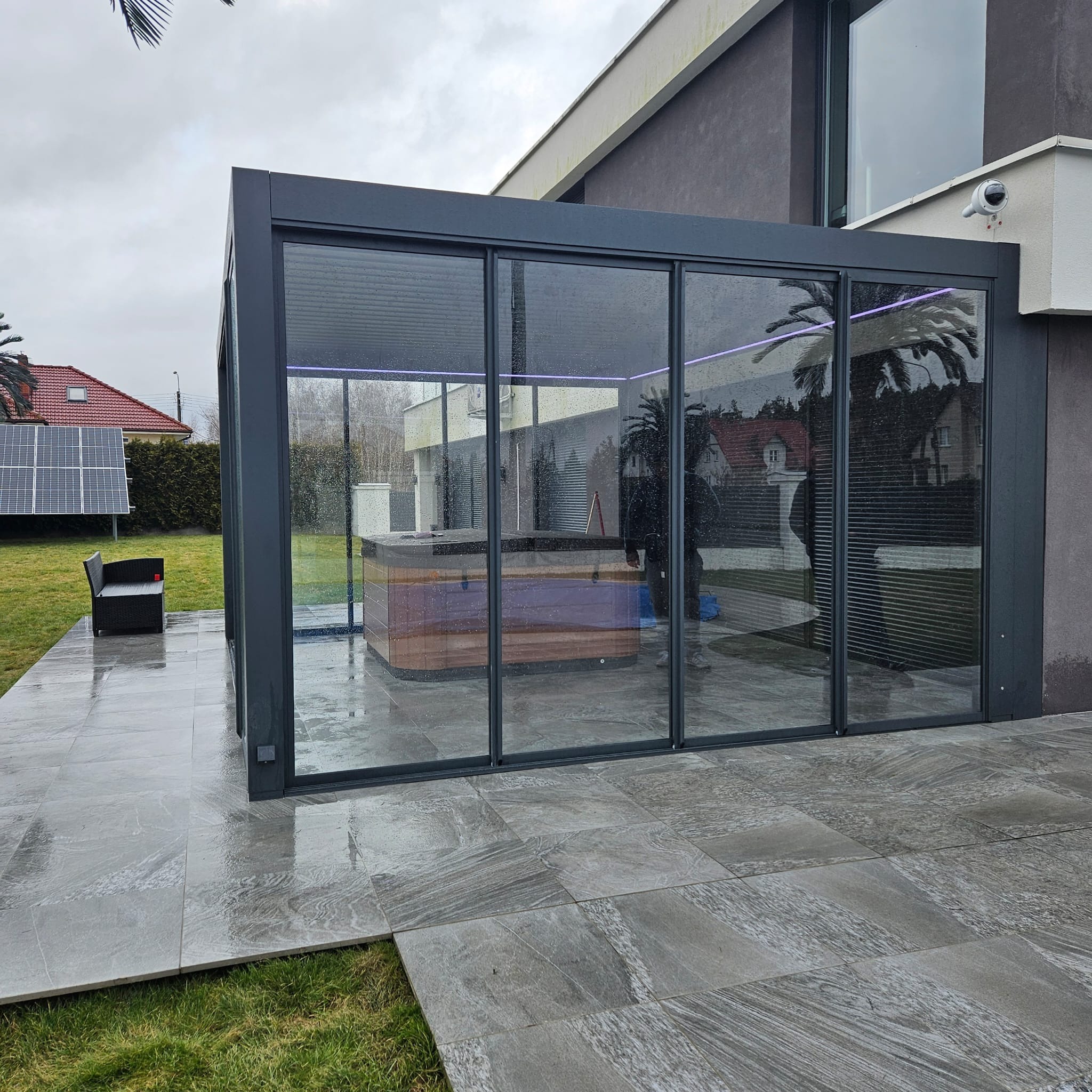 Nowoczesna pergola z szarymi ramami i szklanymi ścianami, z jacuzzi w środku. Na zewnątrz szara kostka brukowa, trawnik i panel słoneczny. Widok na dom.