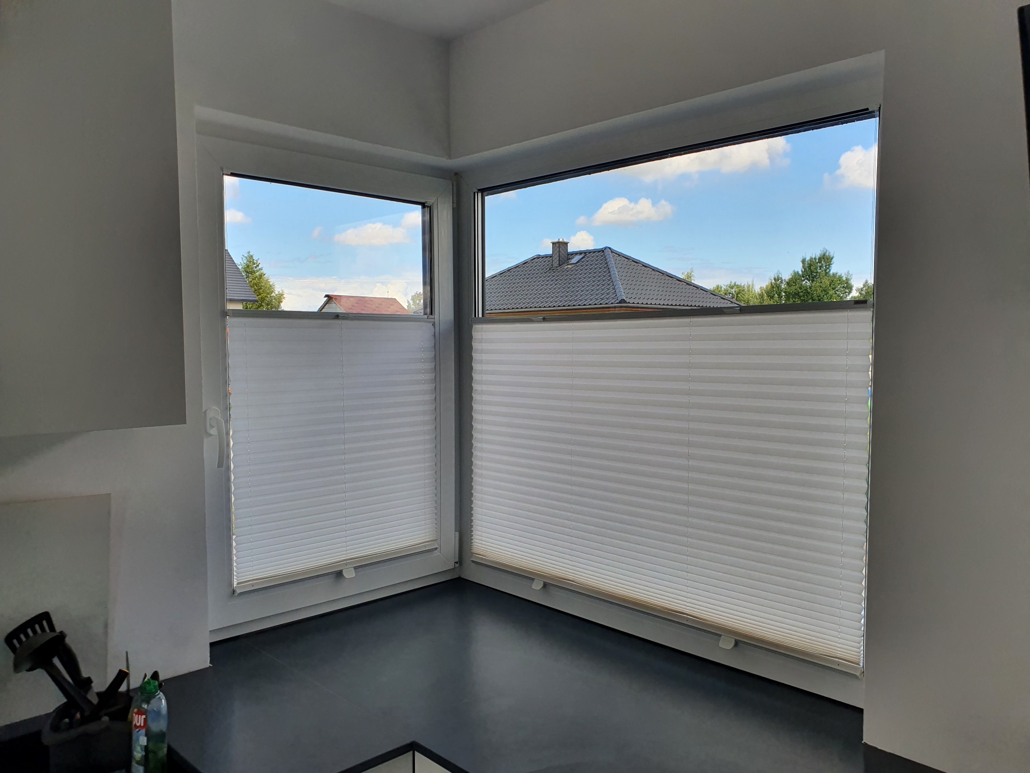 Dwa okna narożne z białymi, plisowanymi osłonami, przez które widać fragment dachu i błękitne niebo. Czarny blat pod oknami i białe ściany.