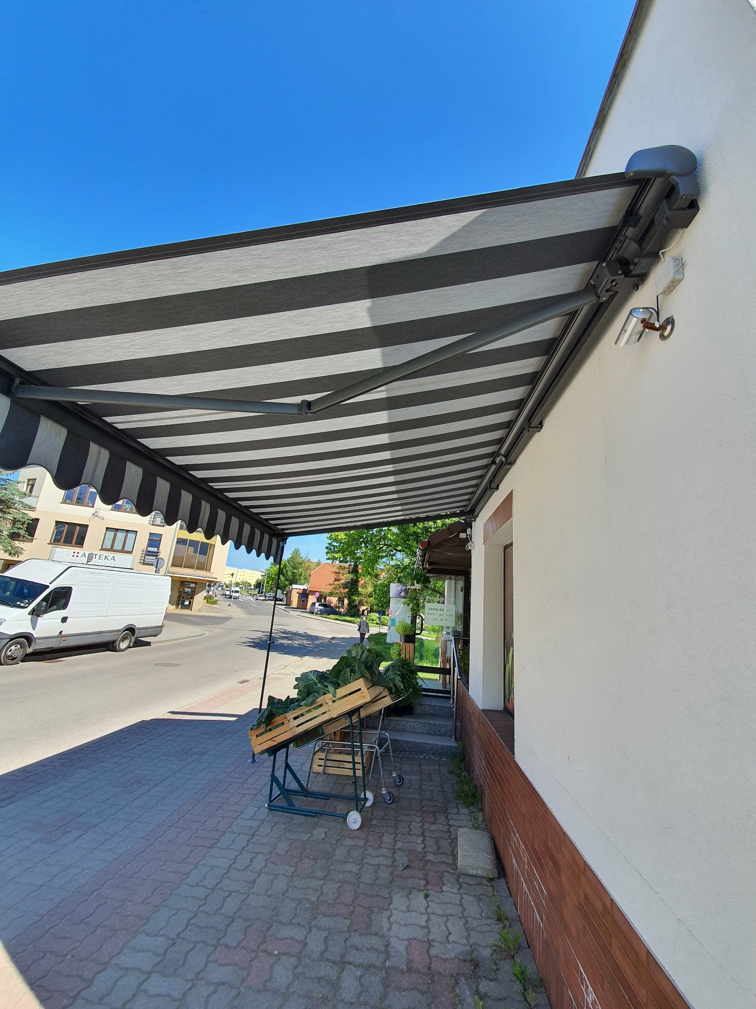 Zewnętrzna, zwijana markiza w czarno-białe pasy, zamontowana nad chodnikiem przy budynku. Widok na ulicę z białym vanem w tle.