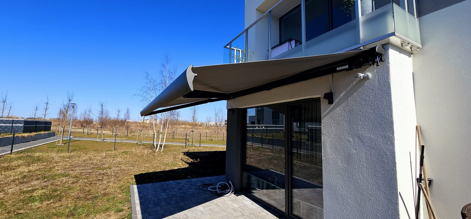 Szara markiza tarasowa zamontowana nad szklanymi drzwiami balkonowymi w nowoczesnym domu z jasną elewacją. Widok na zielony teren i błękitne niebo.