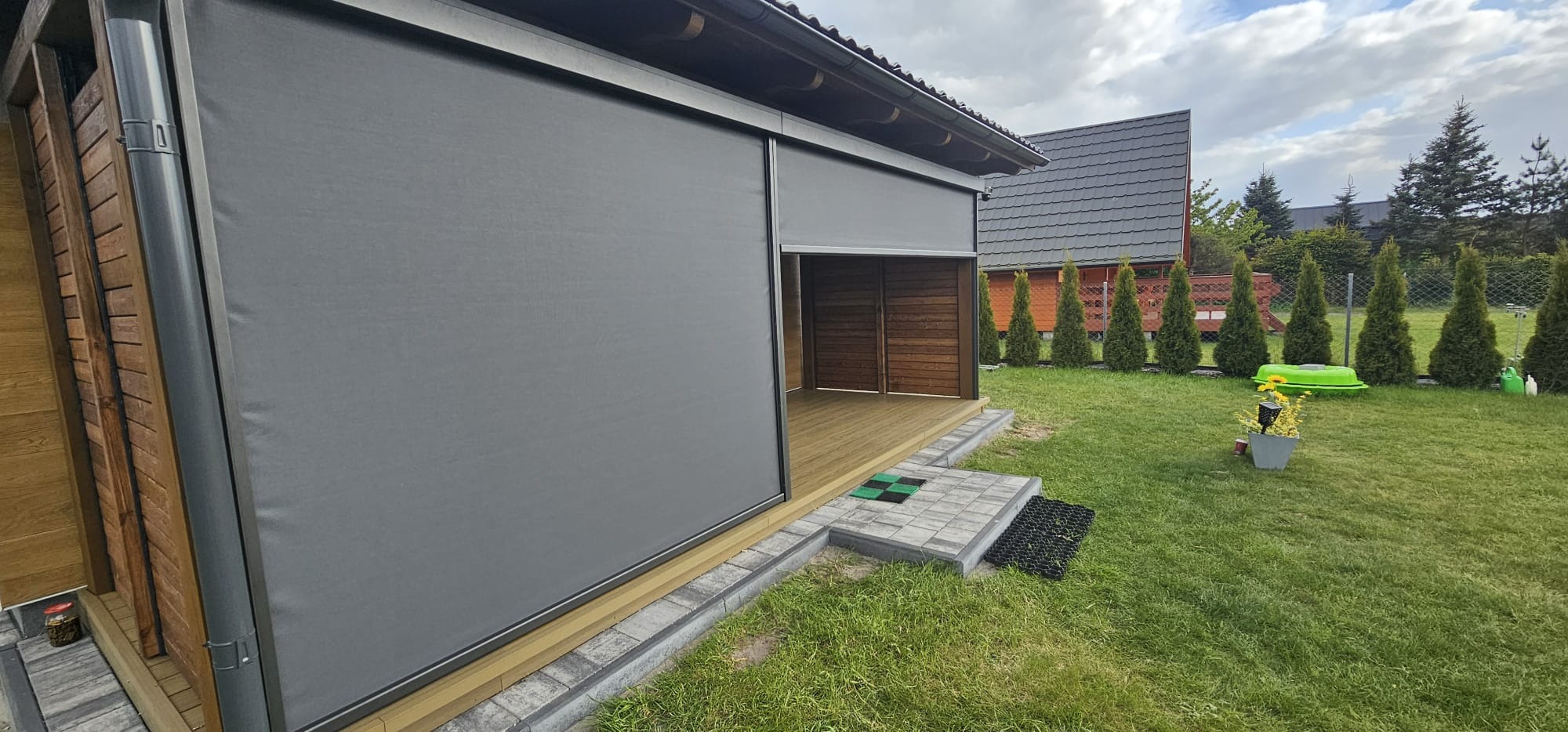 Szara, zwijana przesłona tarasowa częściowo opuszczona, widok na drewniany taras i fragment ogrodu z tujami. Zewnętrzna osłona przeciwsłoneczna.