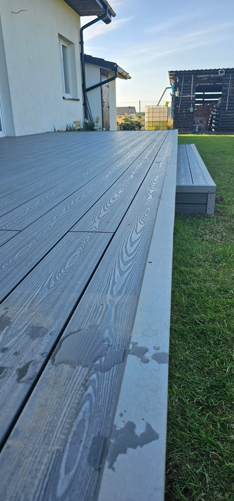 Szary, kompozytowy taras z widocznymi śladami wody, przylegający do białego budynku z zielonym trawnikiem w tle. Ujęcie z niskiego kąta podkreśla strukturę desek.