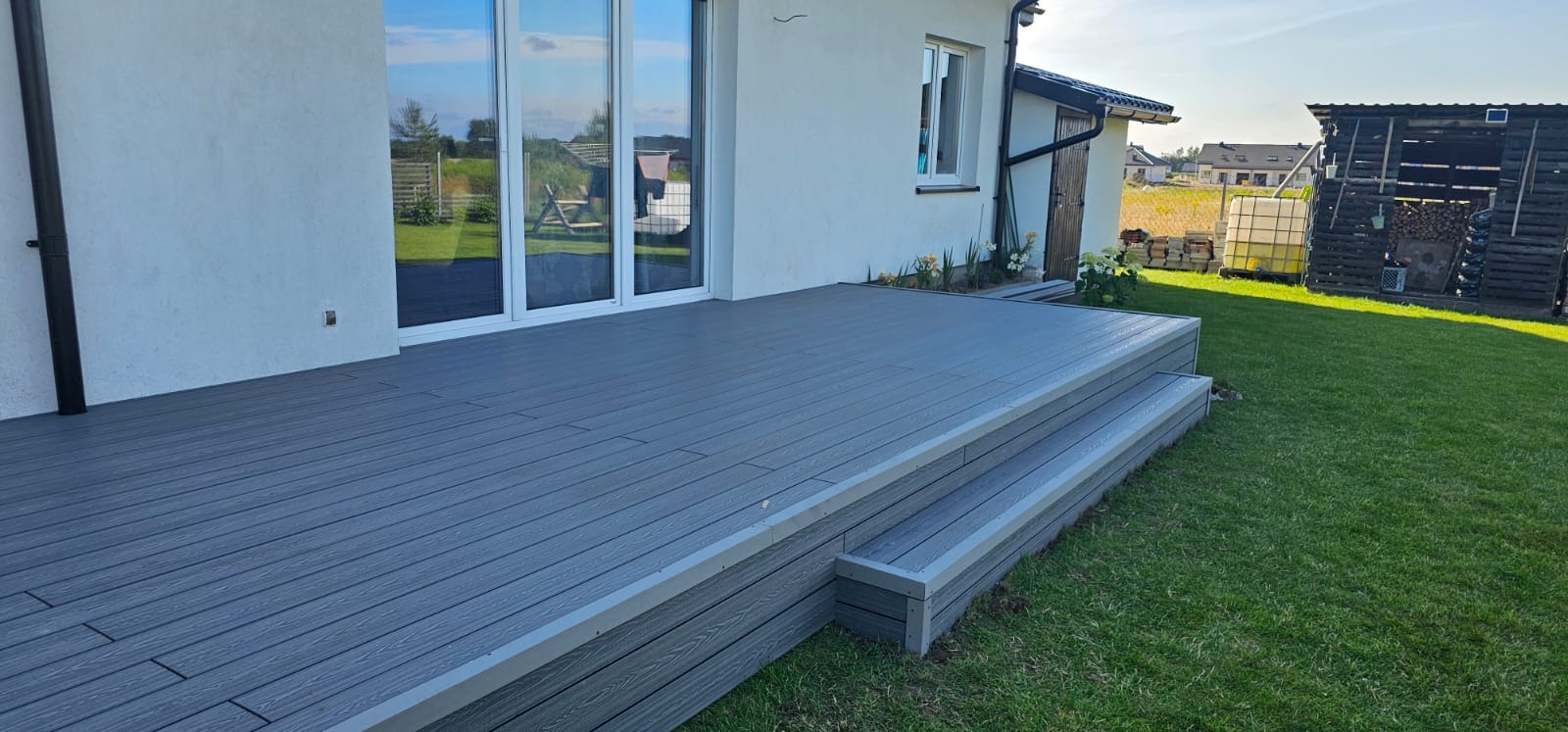 Szara, deska kompozytowa na tarasie z niskimi schodami, na tle domu z białymi ścianami i zielonym trawnikiem. Widoczne odbicia w oknach.