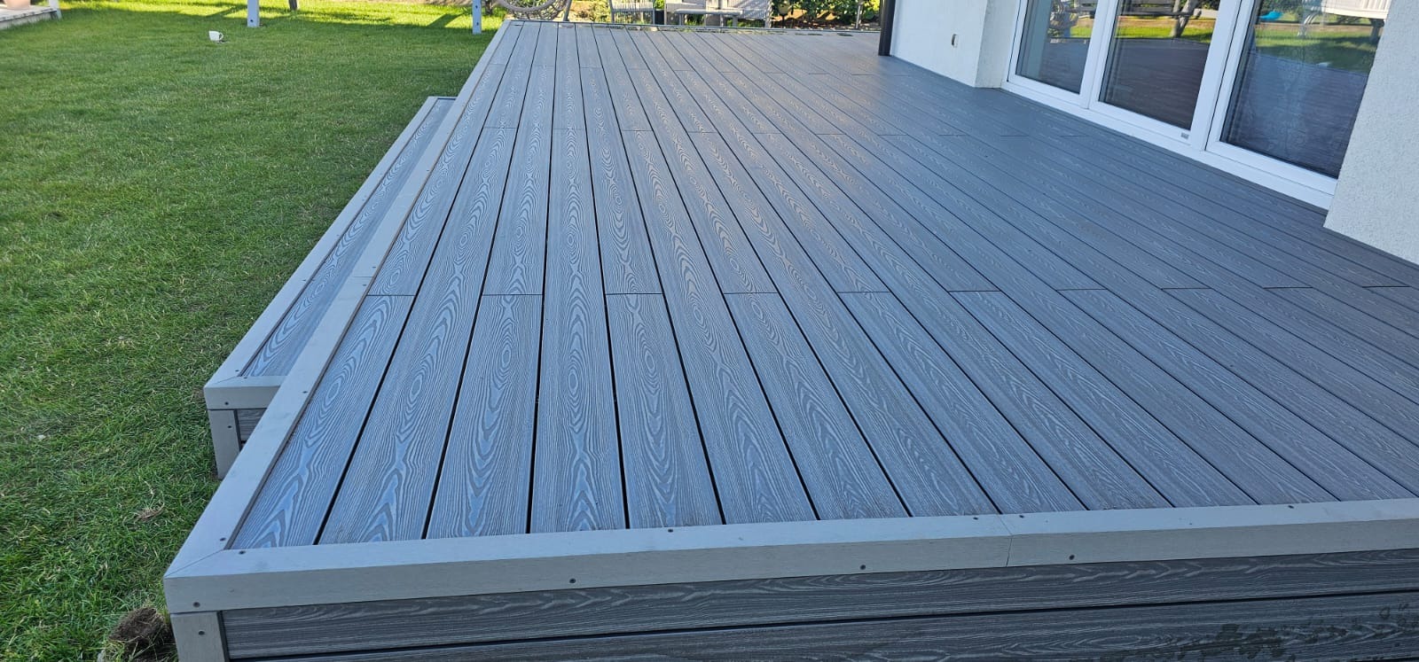 Szary taras kompozytowy z subtelnym wzorem drewna, prowadzący do białych drzwi balkonowych i otoczony zielonym trawnikiem w słoneczny dzień.