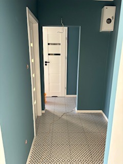 Korytarz po remoncie: ściany pomalowane na ciemny, morski kolor, białe drzwi z przeszkleniami, wzorzysta, czarno-biała podłoga z płytek. Na ścianie widoczna biała skrzynka elektryczna.