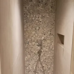 Wykończona kabina prysznicowa z mozaiką z drobnych kamieni w odcieniach szarości i beżu, srebrna armatura i odpływ liniowy, wnęka w ścianie na kosmetyki.