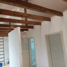 Wnętrze po remoncie: bielone ściany, surowe drewniane belki stropowe, dwoje drzwi wewnętrznych, widoczna łazienka z kabiną prysznicową w tle, drewniana podłoga, drabina oparta o ścianę z białymi...