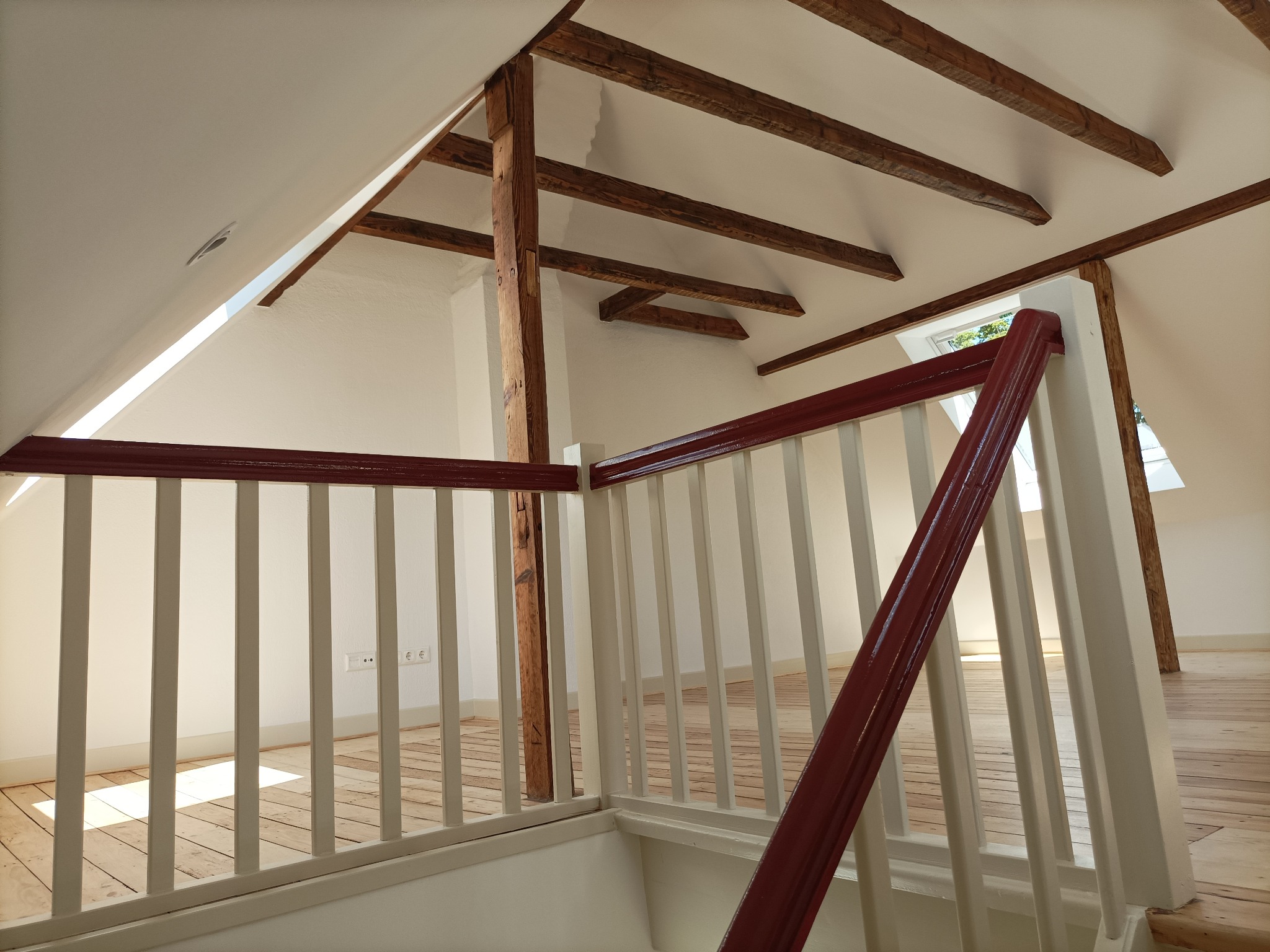 Poddasze po remoncie: biel ścian, drewniane belki stropowe i podłoga, biało-czerwona balustrada schodów, okno dachowe w skosie.