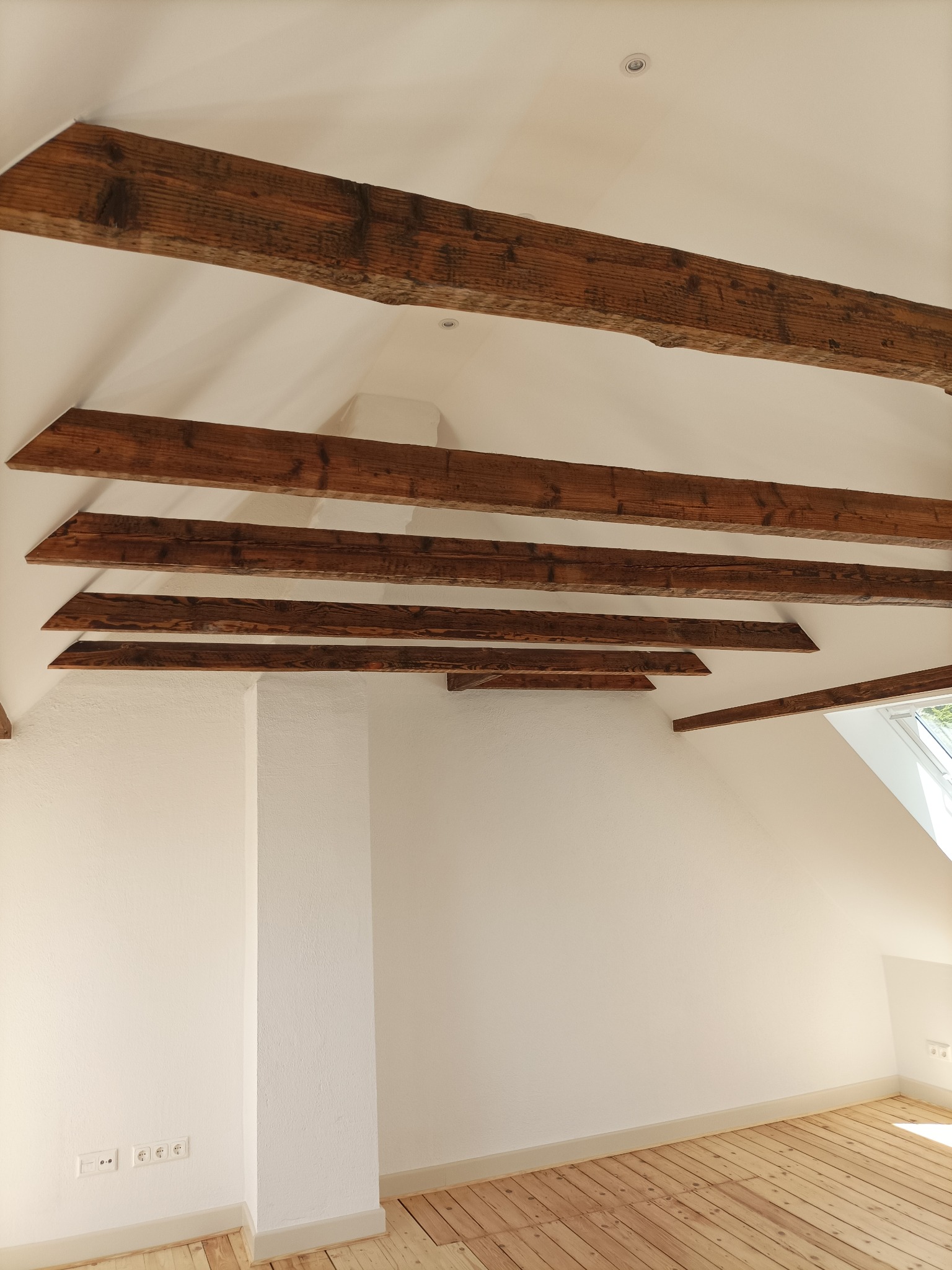 Poddasze po remoncie: świeżo pomalowane białe ściany, drewniana podłoga, widoczne drewniane belki stropowe i oświetlenie punktowe.