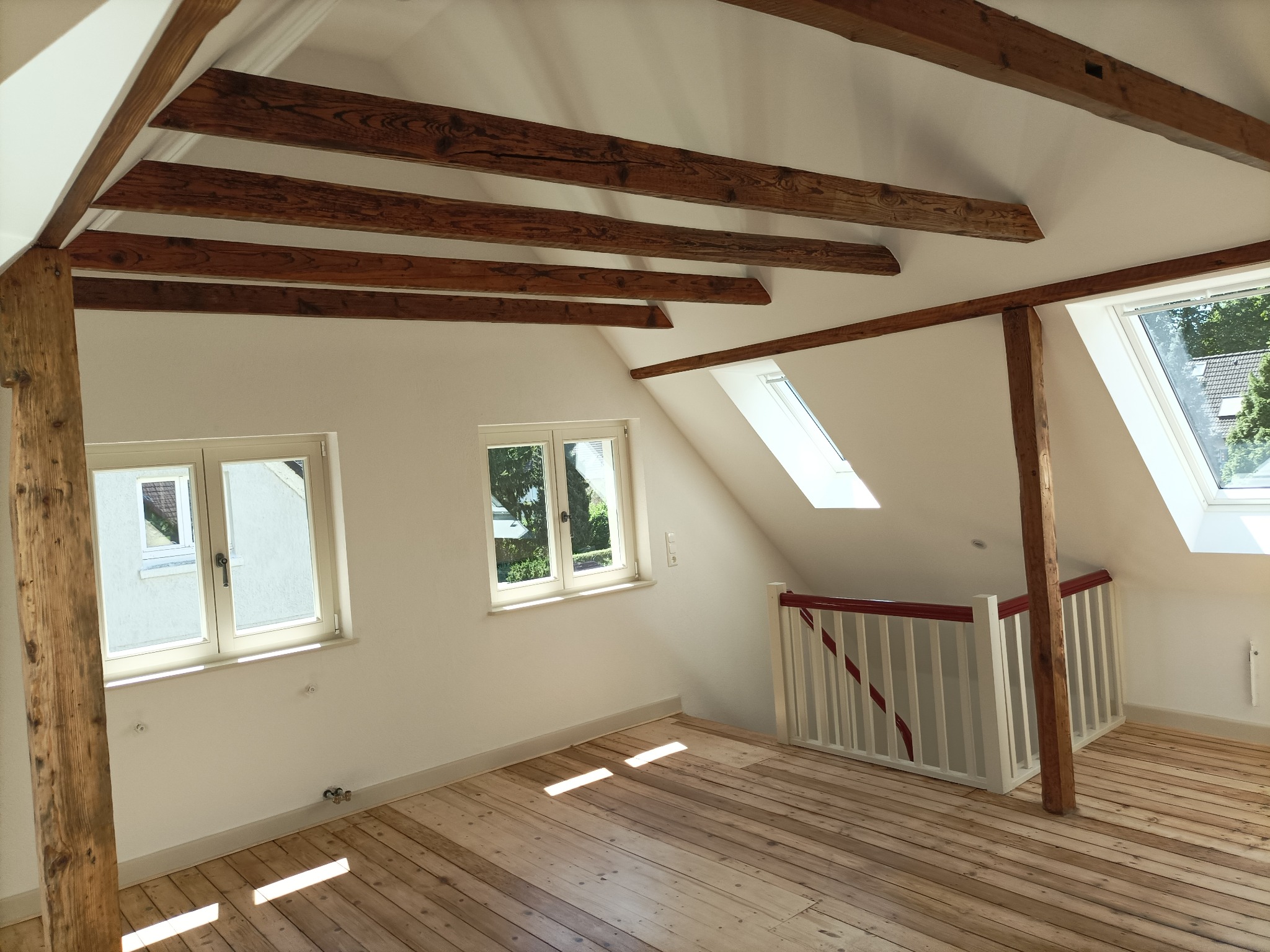 Jasne poddasze z drewnianą podłogą, widoczne belki stropowe i okna dachowe, ściany pomalowane na biało, drewniany słup konstrukcyjny.
