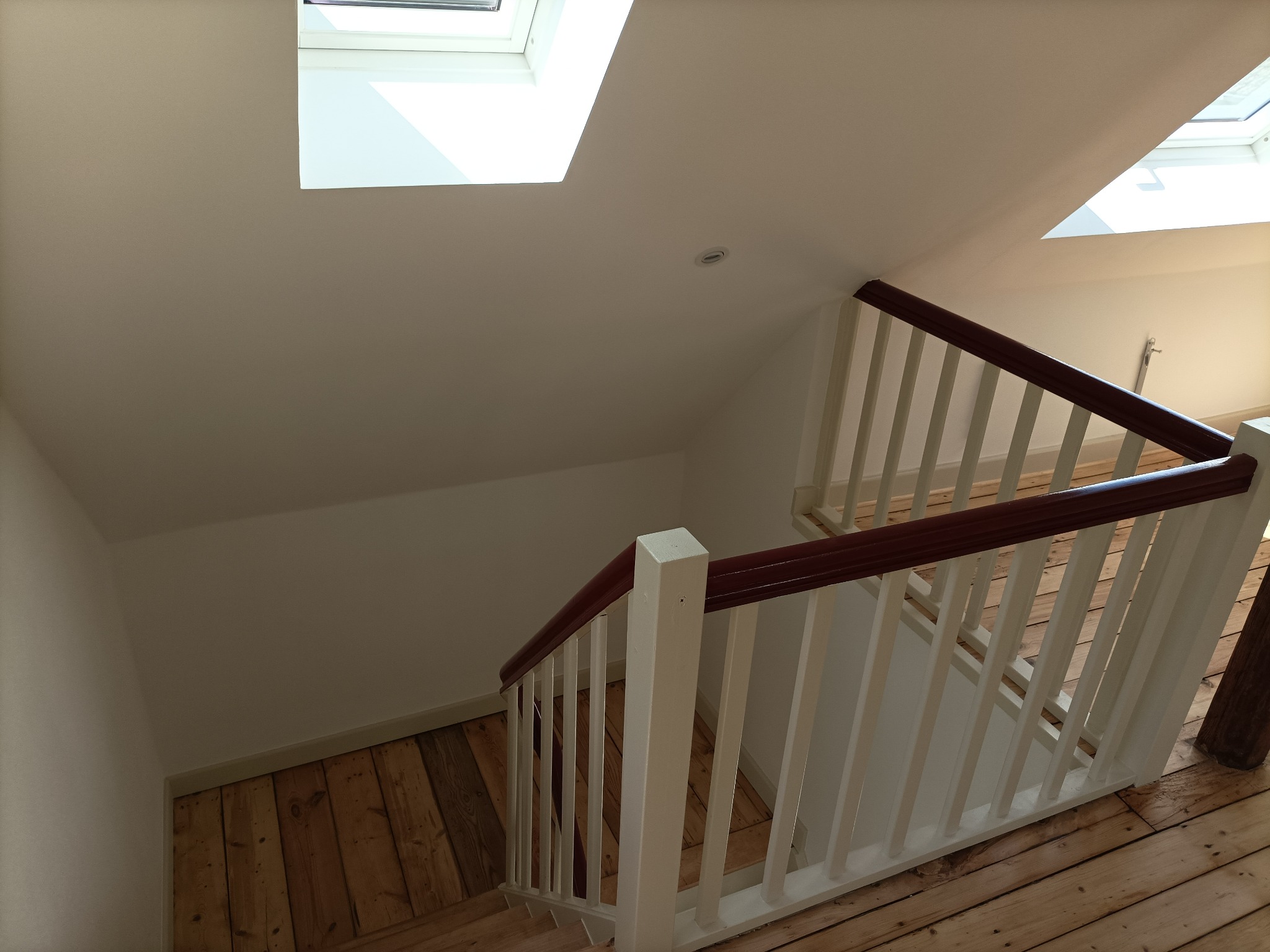 Schody drewniane z białymi balustradami i mahoniową poręczą, widok z góry w jasnym wnętrzu z oknami dachowymi.