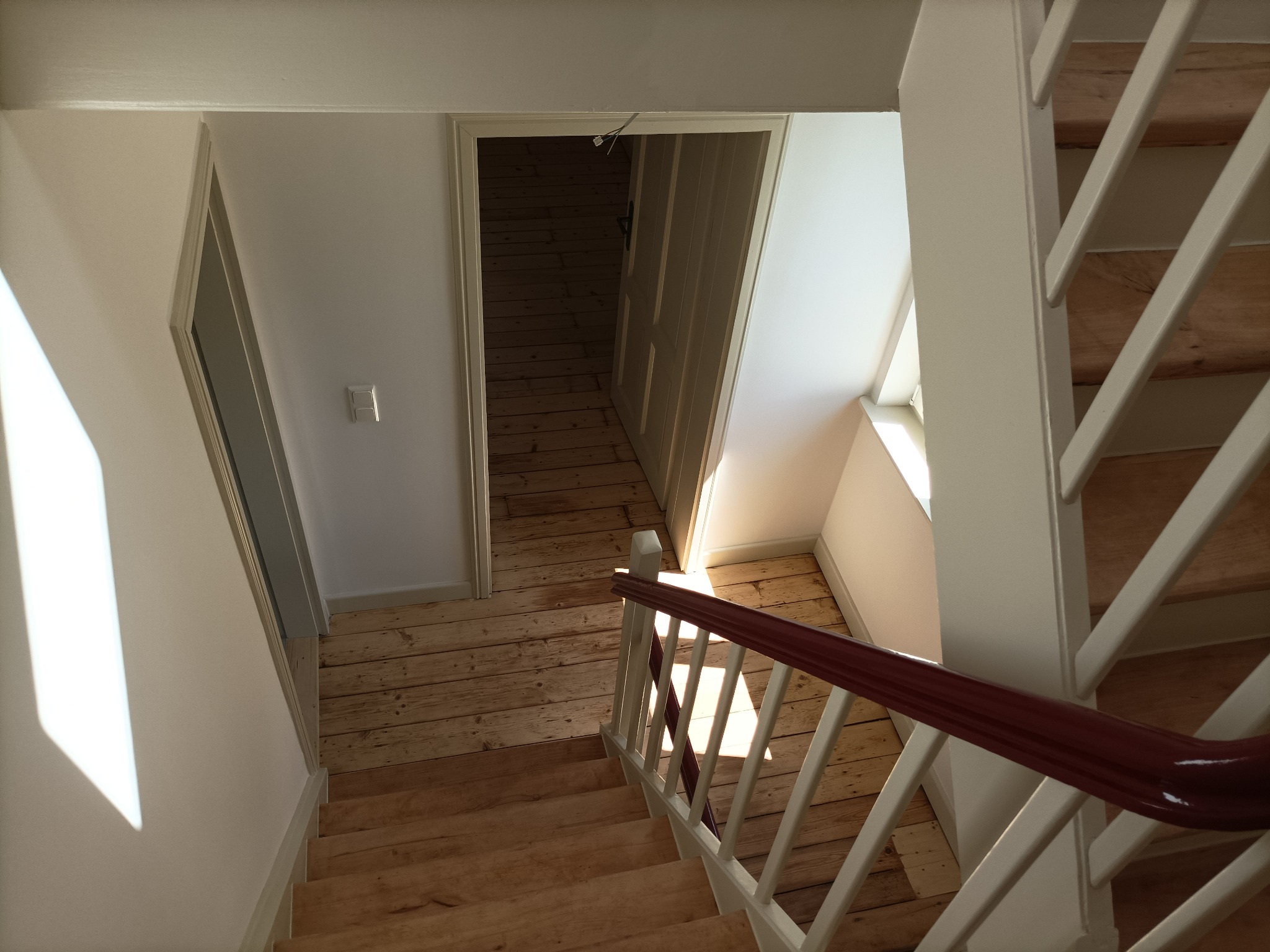 Widok z góry na schody z drewnianymi stopniami i białą balustradą z bordową poręczą, prowadzące w dół do jasnego korytarza z drewnianą podłogą i otwartymi drzwiami do ciemniejszego pomieszczenia.