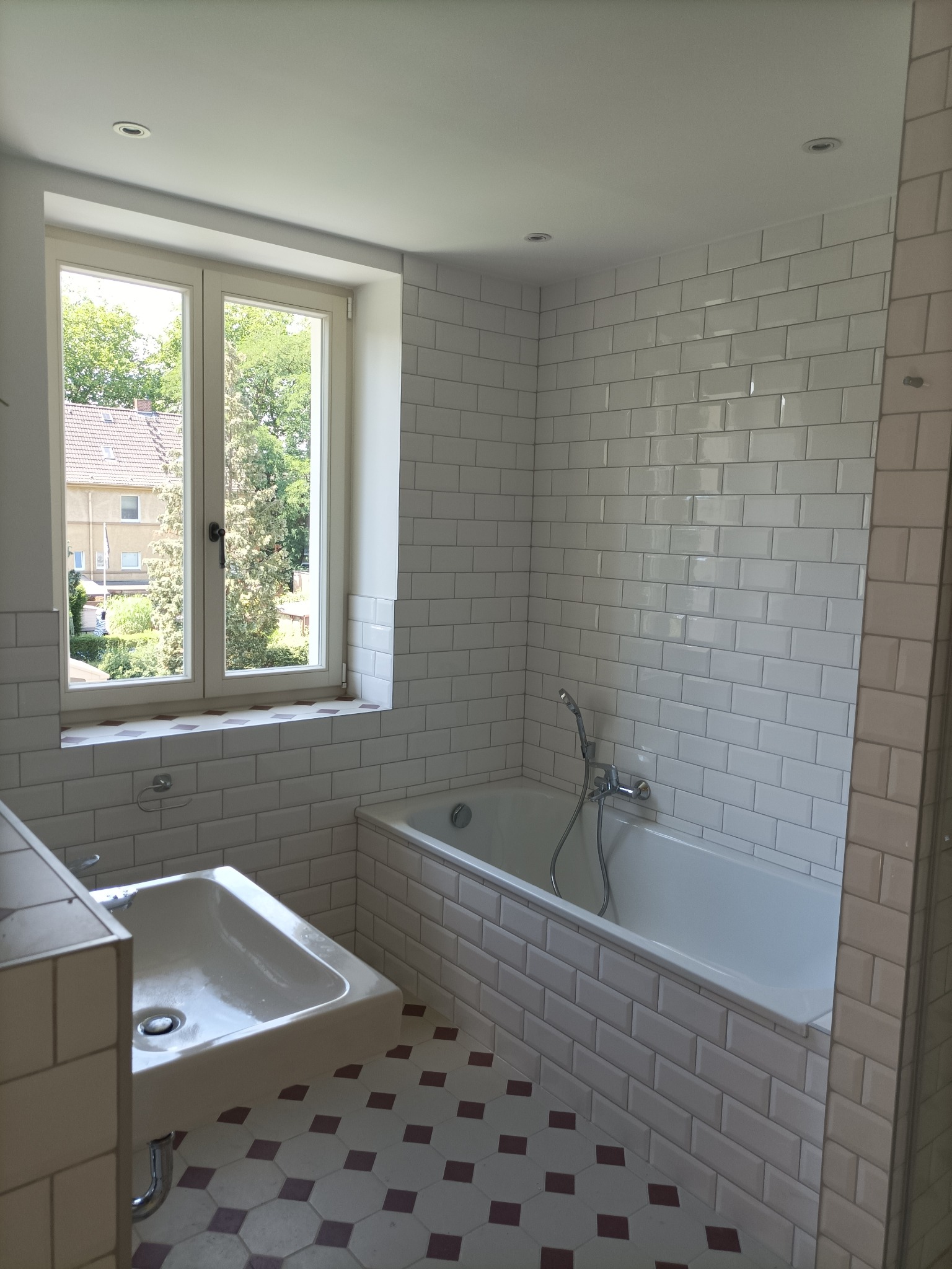 Wnętrze łazienki po remoncie: białe, błyszczące płytki typu metro na ścianach, biała wanna, umywalka z chromowaną baterią, podłoga z heksagonalnych płytek w kolorze kremowym i bordowym, okno...