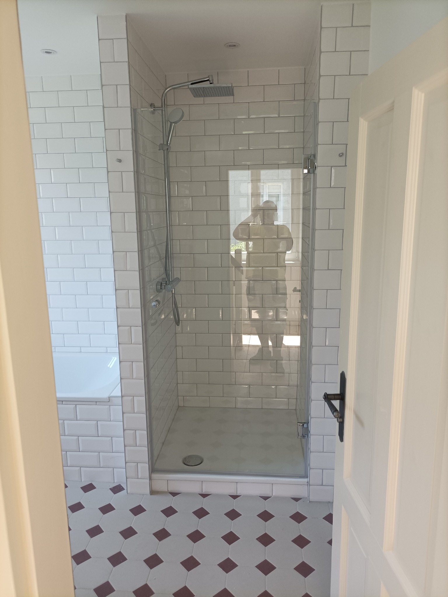 Wykończona łazienka z kabiną prysznicową typu walk-in, białe kafelki typu metro na ścianach, wzorzysta podłoga w odcieniach bieli i bordo, widoczna część wanny i otwarte drzwi z czarną klamką.