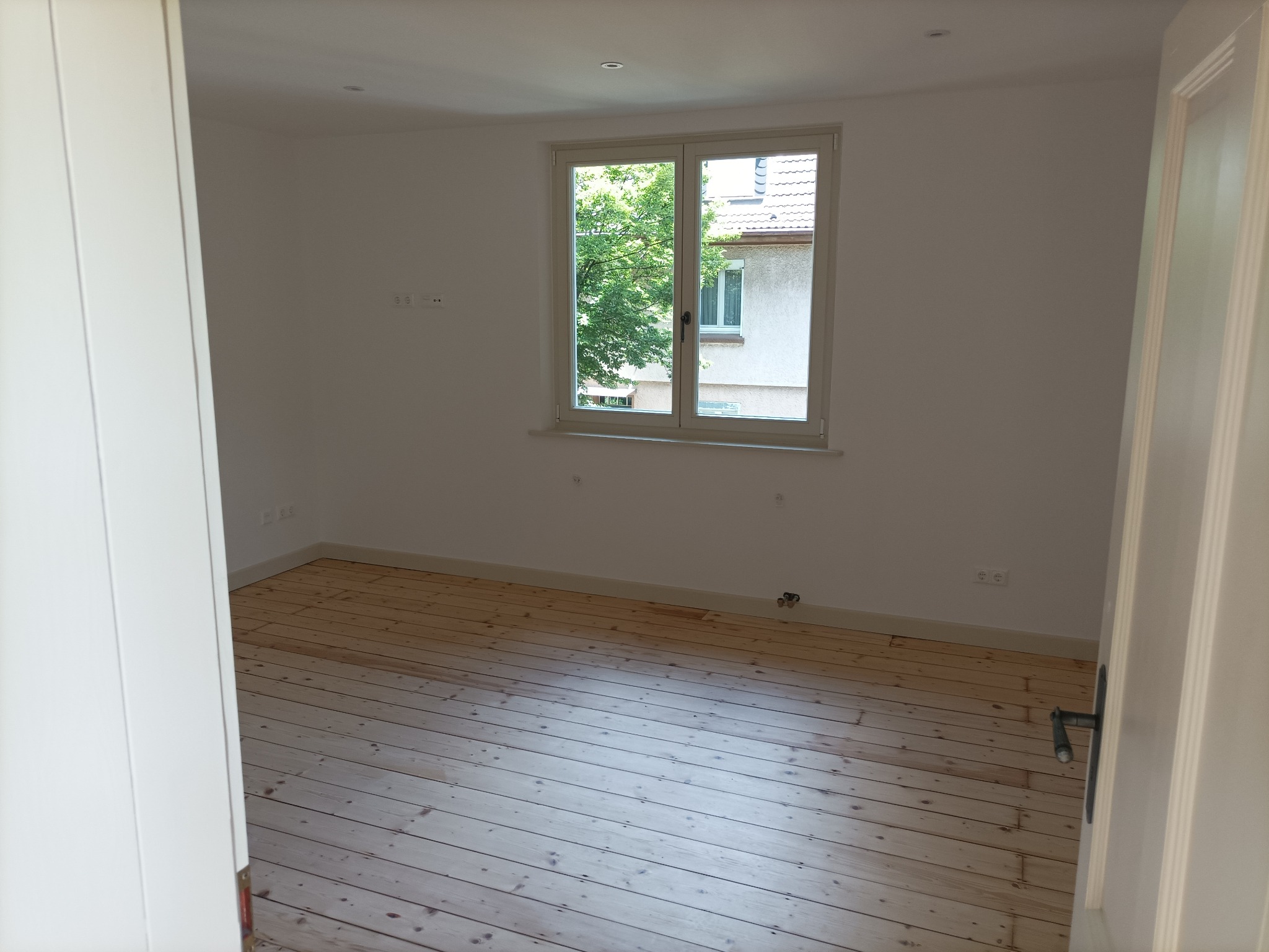 Pusty pokój z jasnymi ścianami, drewnianą podłogą i oknem z widokiem na zieleń, uchwycony z perspektywy otwartych drzwi.