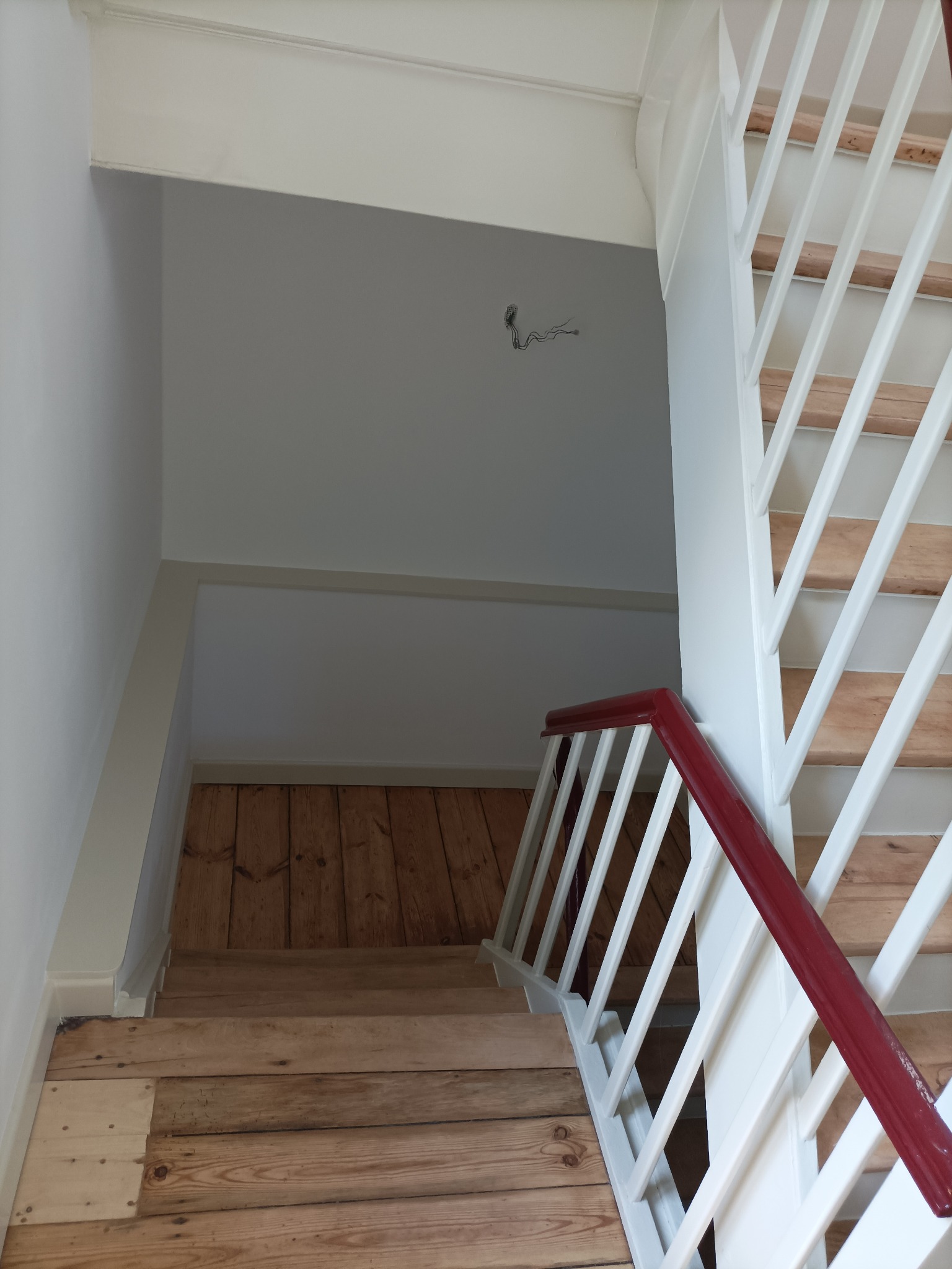 Schody drewniane z pomalowanymi na biało balustradami i bordową poręczą, ściany w odcieniach bieli i szarości, widoczne przewody elektryczne.