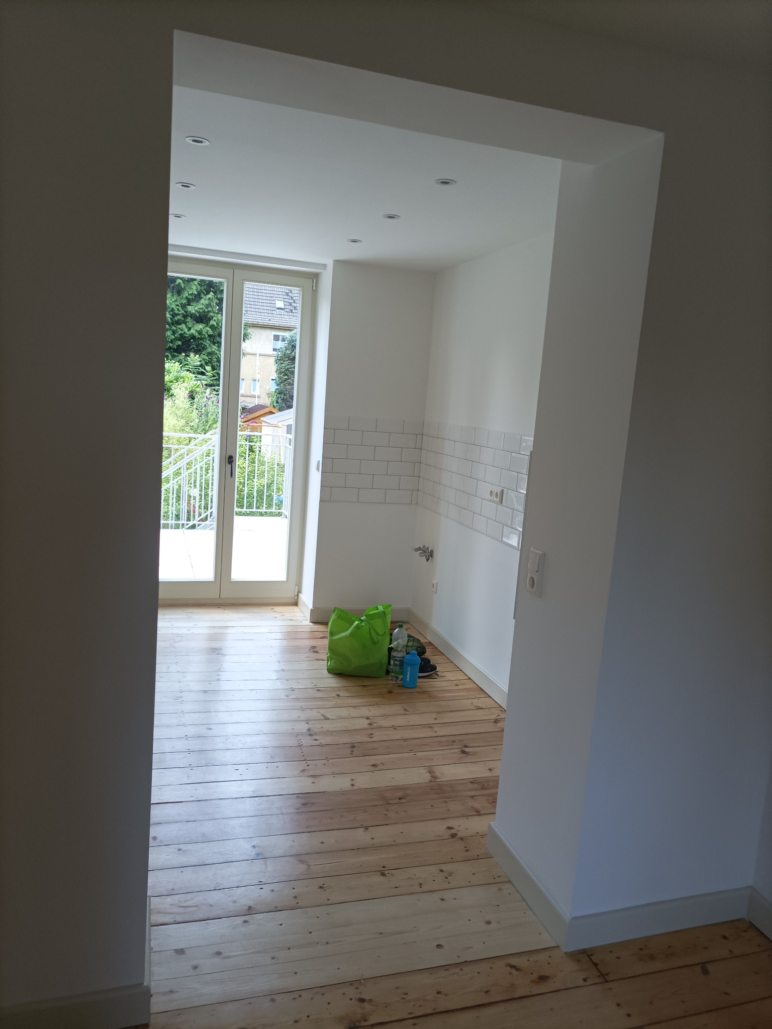 Wnętrze wykończonego pokoju z drewnianą podłogą, białymi ścianami z fragmentem białych płytek na ścianie, widok na balkon przez otwarte drzwi balkonowe, zielona torba na podłodze.