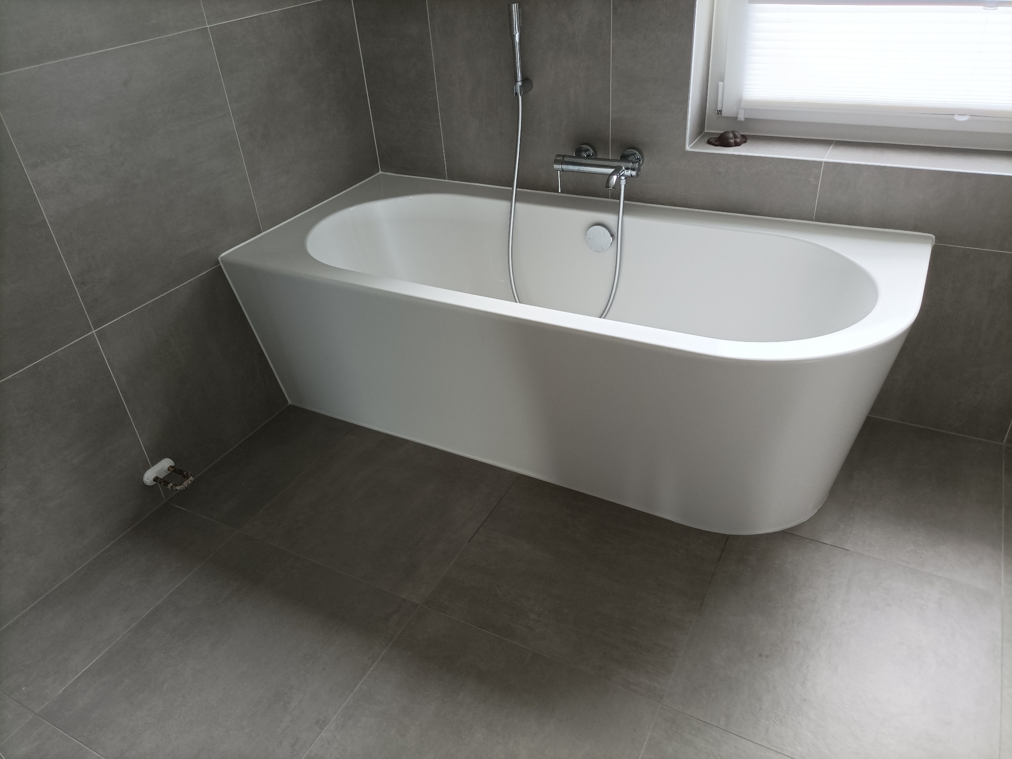 Nowoczesna łazienka z wanną w zabudowie, szare płytki na ścianach i podłodze, chromowana armatura, okno z żaluzjami.