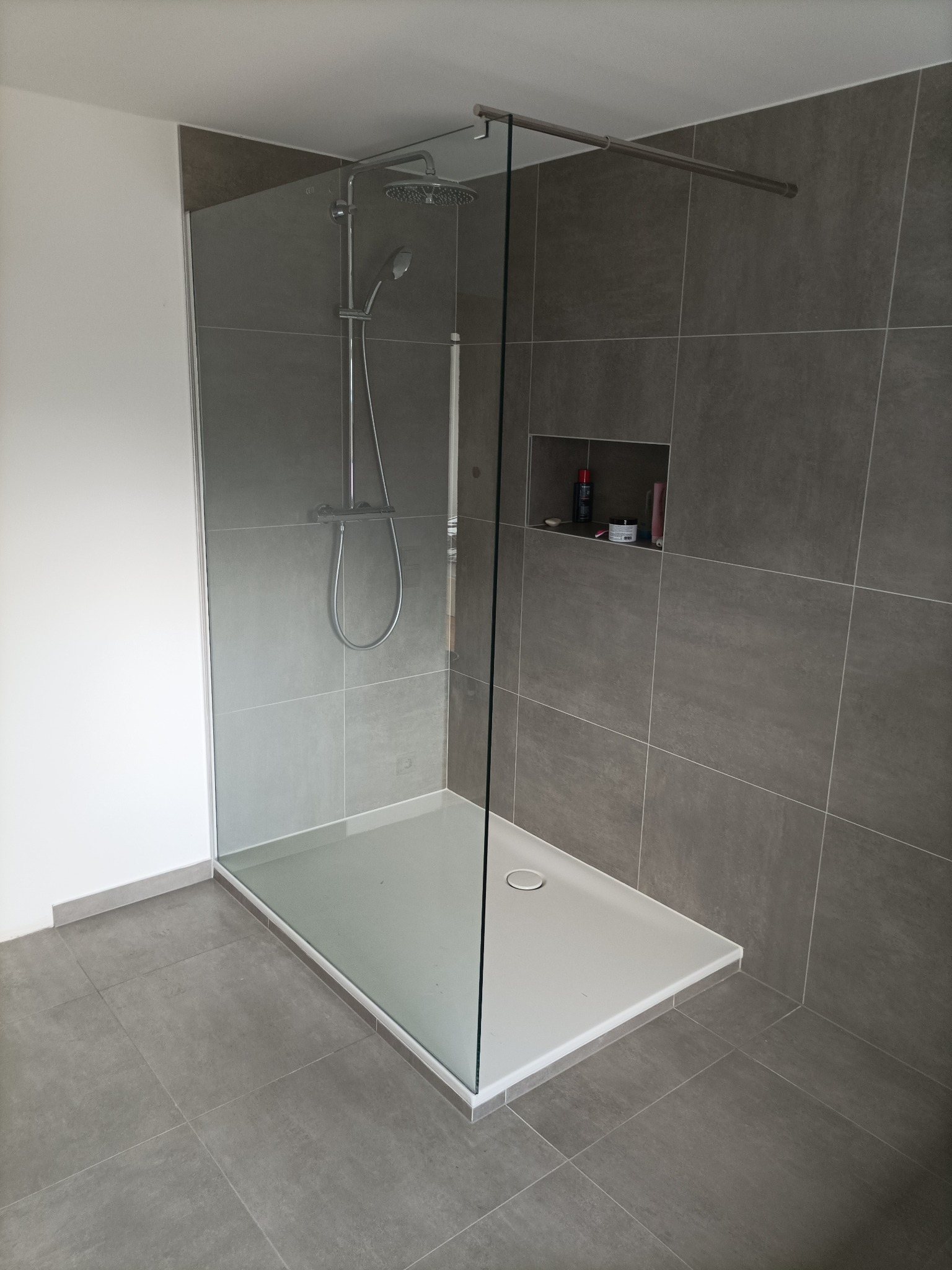 Nowoczesna kabina prysznicowa z szarymi płytkami, szklaną ścianką i białą brodzikiem; wnęka w ścianie z kosmetykami.