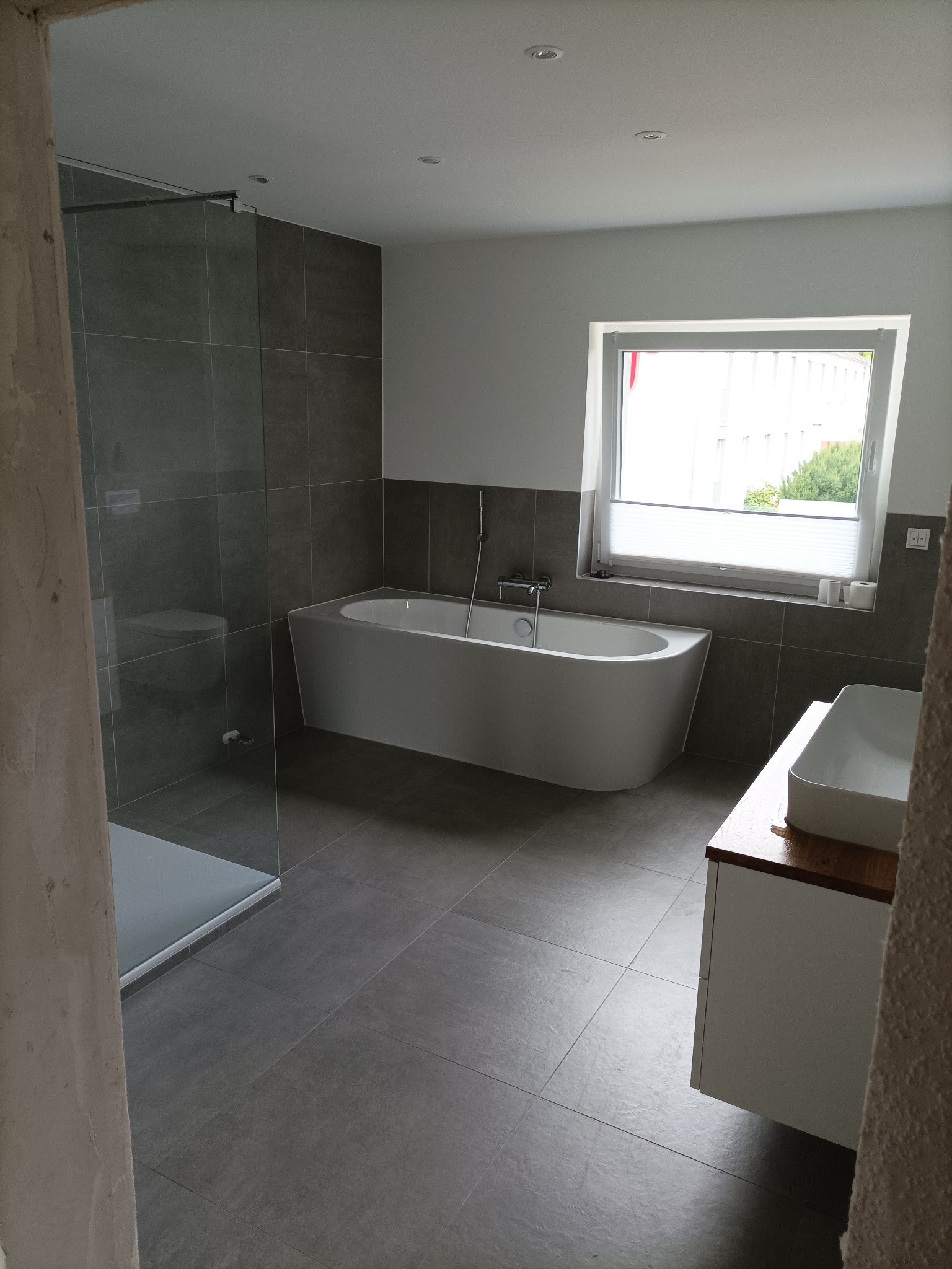 Nowoczesna łazienka z szarymi płytkami, białą wanną wolnostojącą, kabiną prysznicową typu walk-in i umywalką nablatową na szafce z drewnianym blatem, widok zza surowego fragmentu ściany.