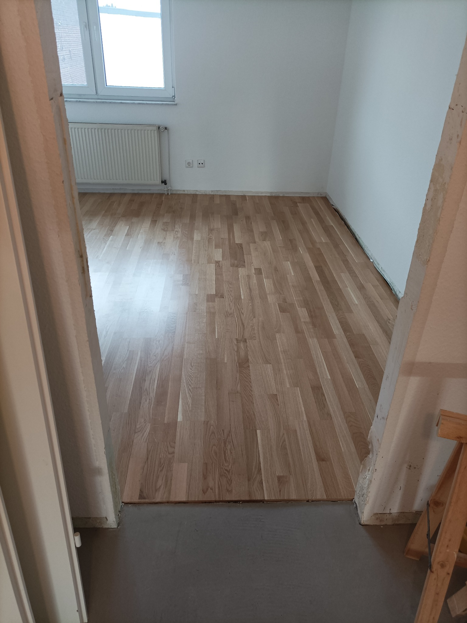 Jasne panele podłogowe ułożone w pustym pokoju z białymi ścianami, widok przez framugę drzwi, grzejnik pod oknem.