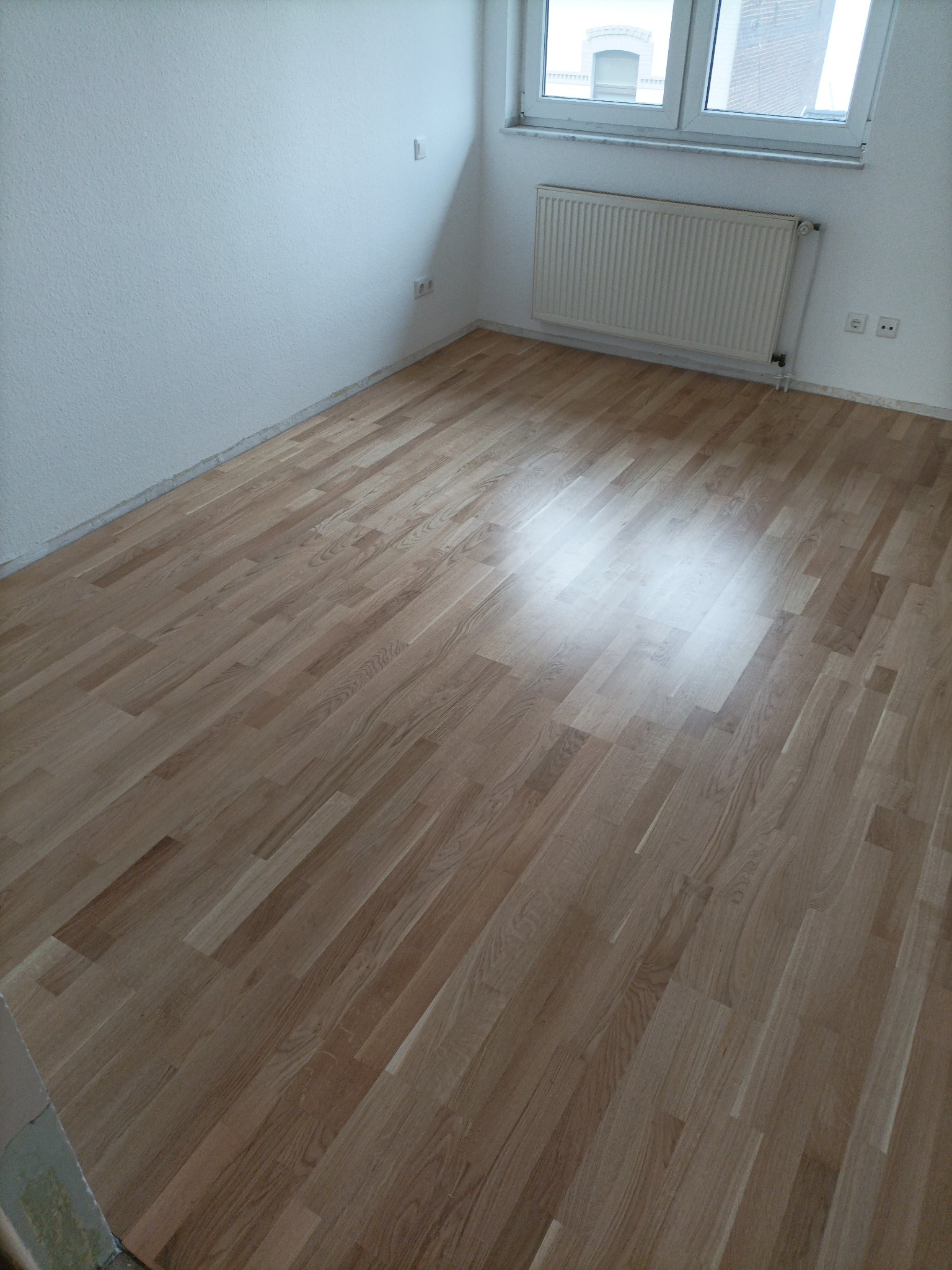 Jasne panele podłogowe ułożone w pokoju z białymi ścianami, grzejnikiem pod oknem i gniazdkami elektrycznymi przy podłodze.