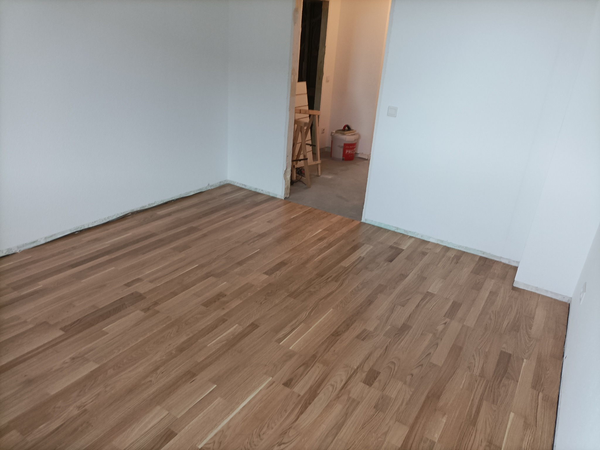 Parkiet ułożony w nowym pomieszczeniu, ściany pomalowane na biało, widoczna drabina malarska i wiadro z farbą w przejściu.