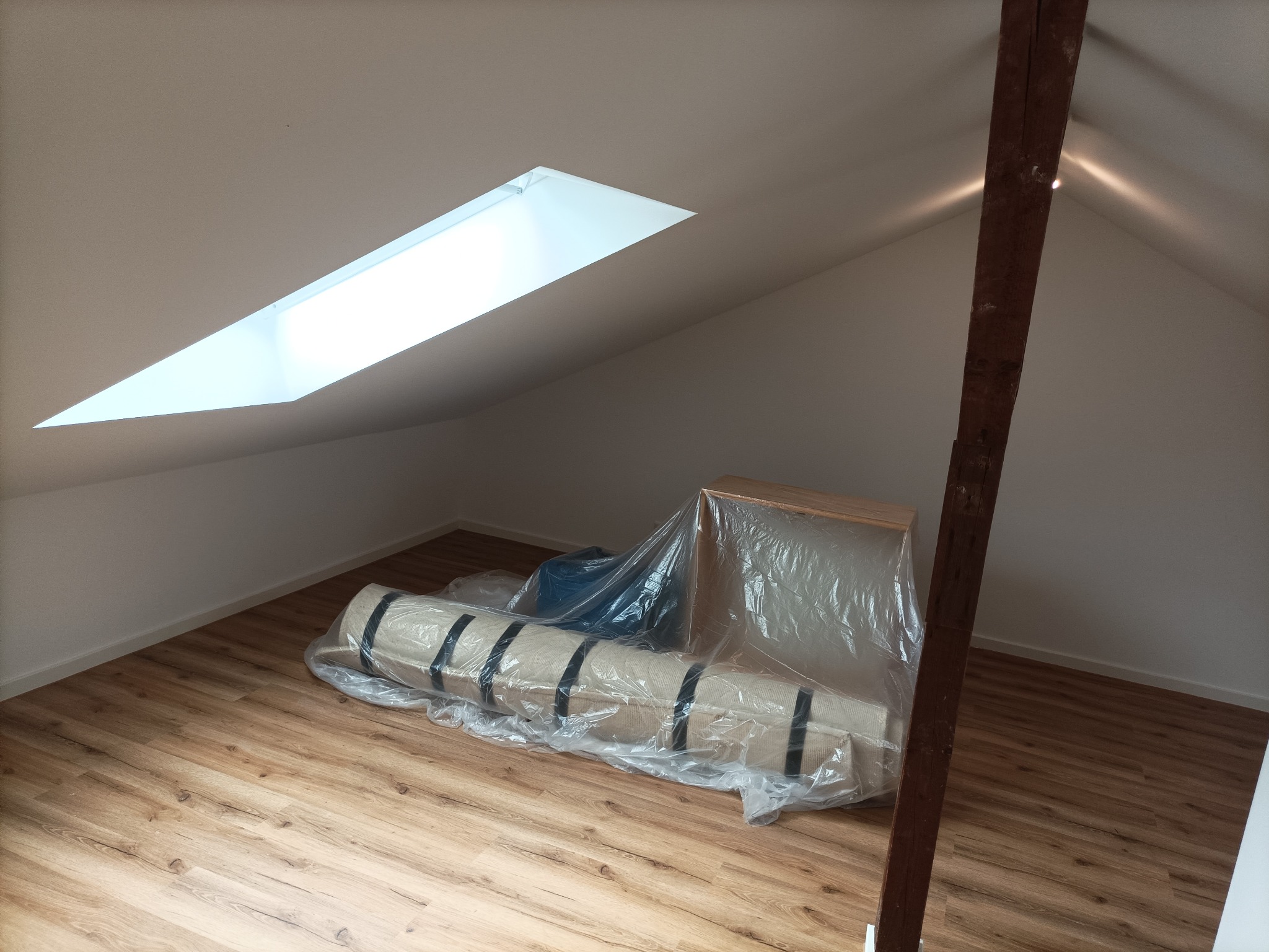 Poddasze z drewnianą podłogą, roletą okienną i belką stropową, przygotowane do prac wykończeniowych; zwinięty dywan i rama okienna zabezpieczone folią.
