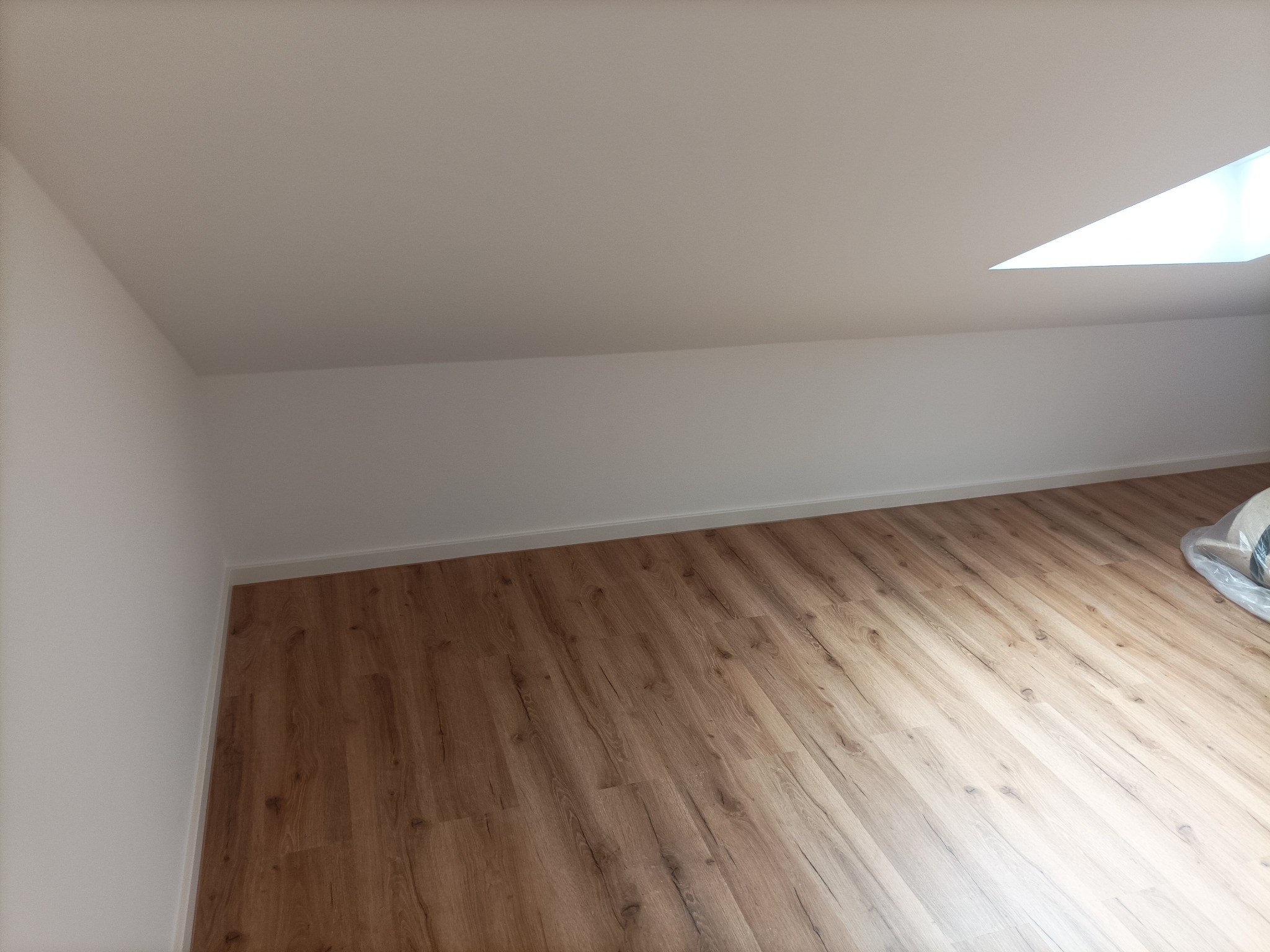 Poddasze po remoncie: Świeżo pomalowane białe ściany, ułożone panele podłogowe z widoczną strukturą drewna, okno dachowe wpuszczające naturalne światło.