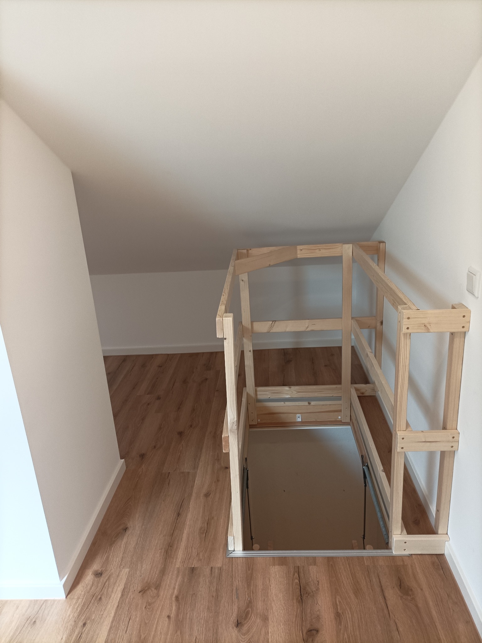 Drewniana konstrukcja zabezpieczająca otwór do piwnicy na poddaszu, widoczne sprężyny mechanizmu otwierania, panele podłogowe i białe ściany.