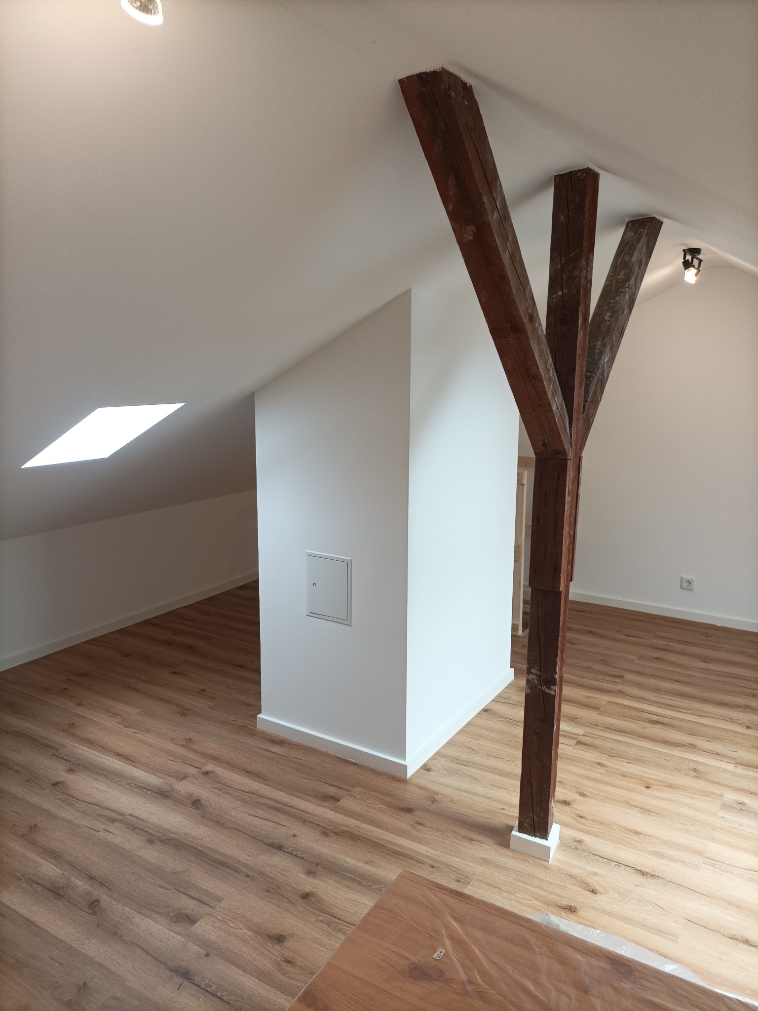 Poddasze po remoncie: świeżo ułożone panele podłogowe w odcieniach brązu, białe ściany z widocznym włazem rewizyjnym, drewniane belki konstrukcyjne w kształcie litery Y, okno dachowe wpadające w skos.