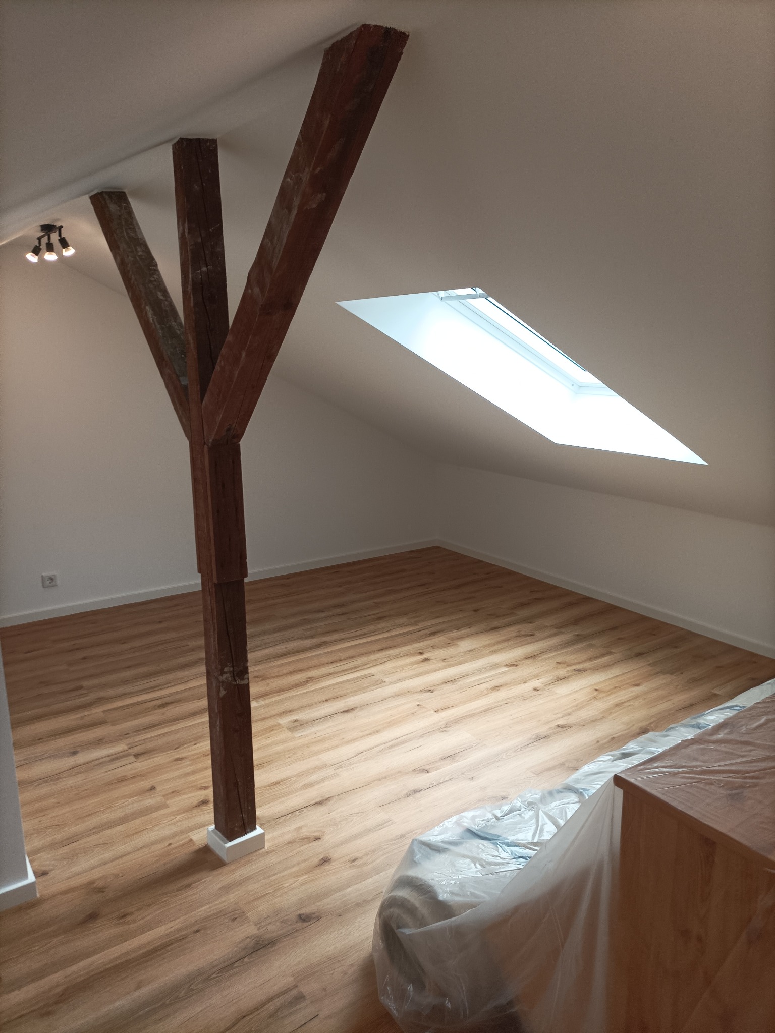 Poddasze po remoncie: świeżo ułożone panele podłogowe w odcieniach drewna, drewniana belka konstrukcyjna, białe ściany i okno dachowe wpadające do pomieszczenia.