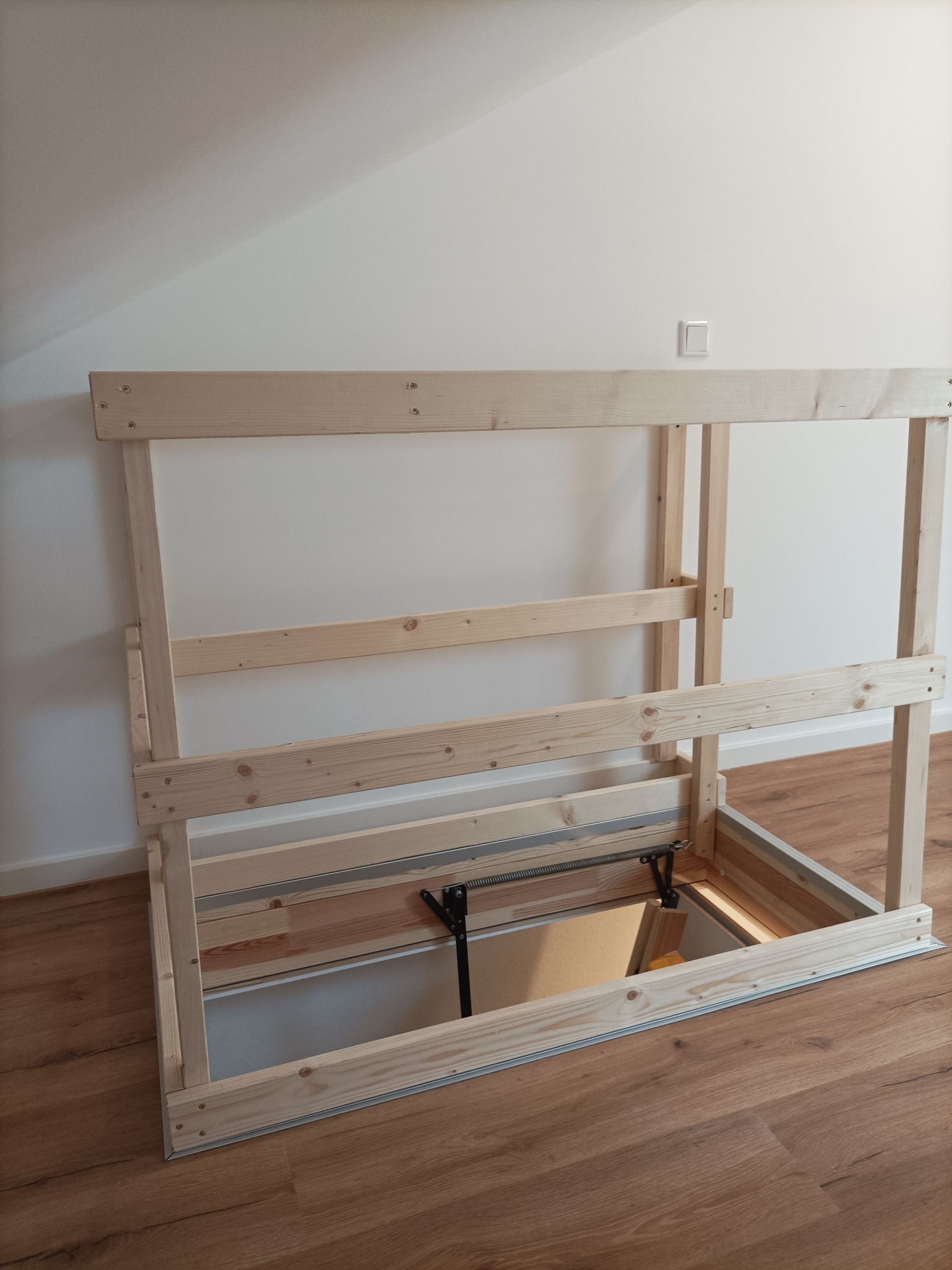 Drewniana rama konstrukcyjna w podłodze z widocznym otworem, mechanizm sprężynowy i metalowe prowadnice, jasne panele podłogowe.