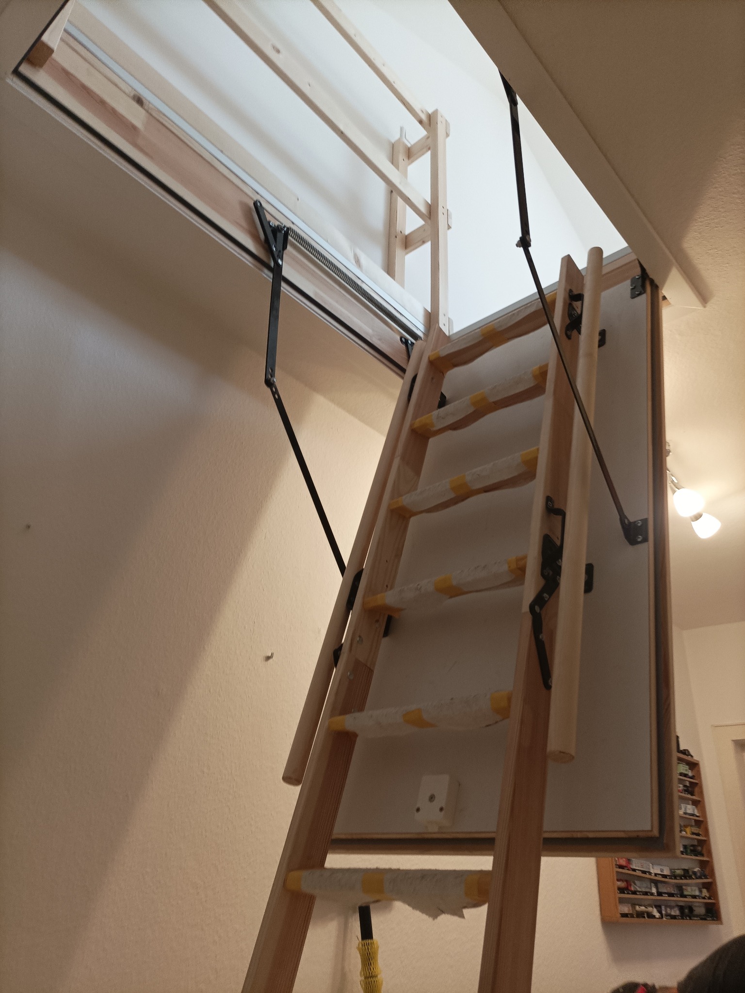 Drewniane schody strychowe, składane, z mechanizmem sprężynowym, częściowo owinięte folią ochronną, widoczne po otwarciu w suficie.