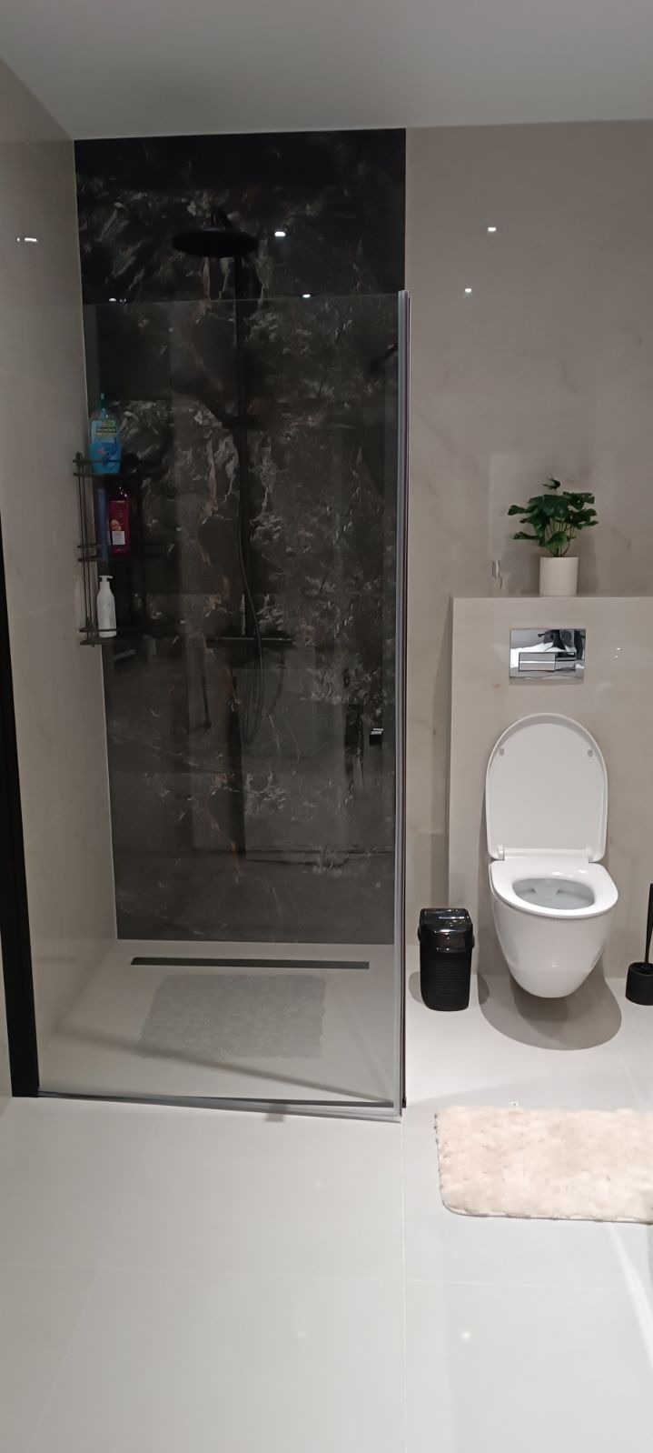 Nowoczesna łazienka z kabiną prysznicową typu walk-in z czarnymi, marmurkowymi płytkami, wiszącą toaletą i minimalistycznymi akcesoriami, takimi jak czarny kosz na śmieci i dywanik z długim włosiem.