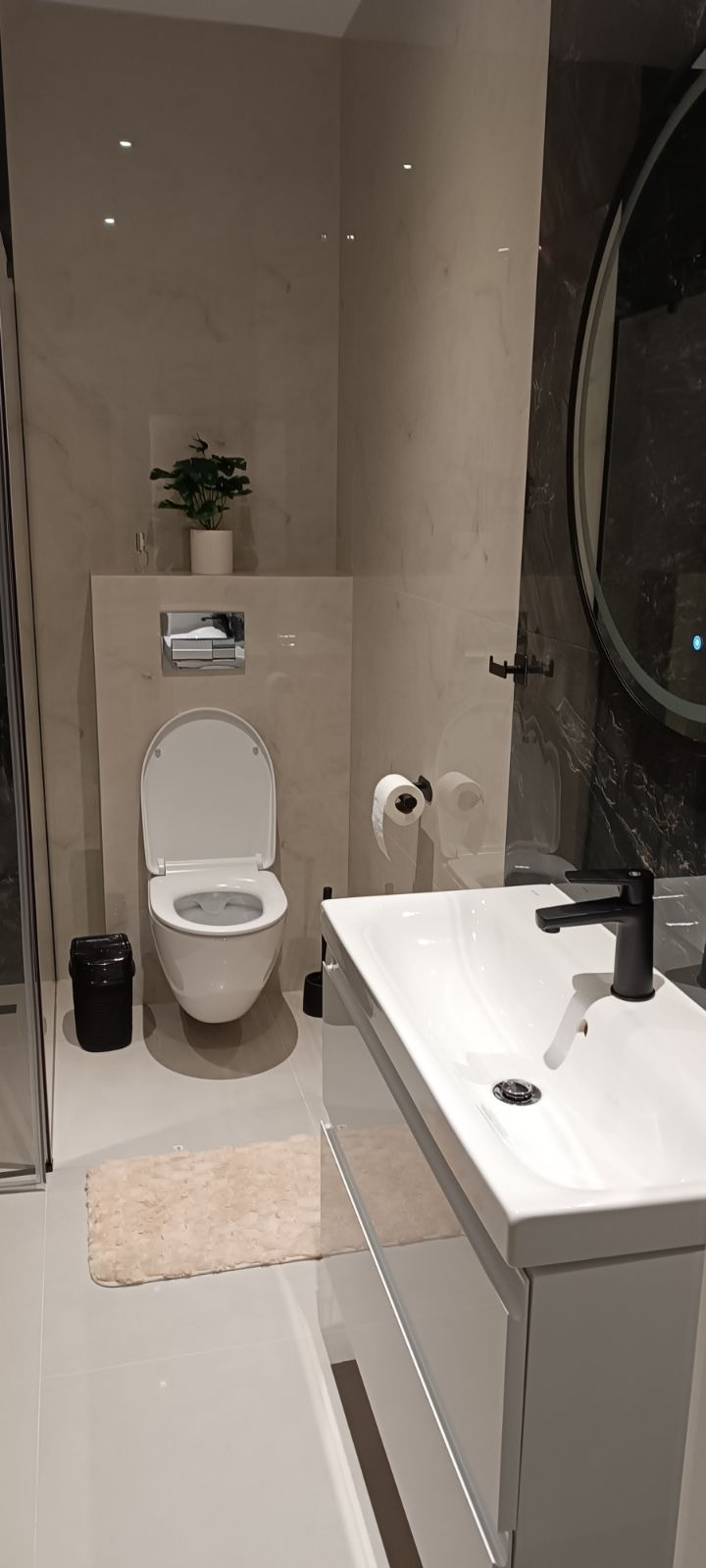 Nowoczesna łazienka w jasnych barwach z wiszącą toaletą, umywalką z czarną armaturą i okrągłym lustrem z podświetleniem LED, uzupełniona o minimalistyczne akcesoria i roślinę doniczkową.