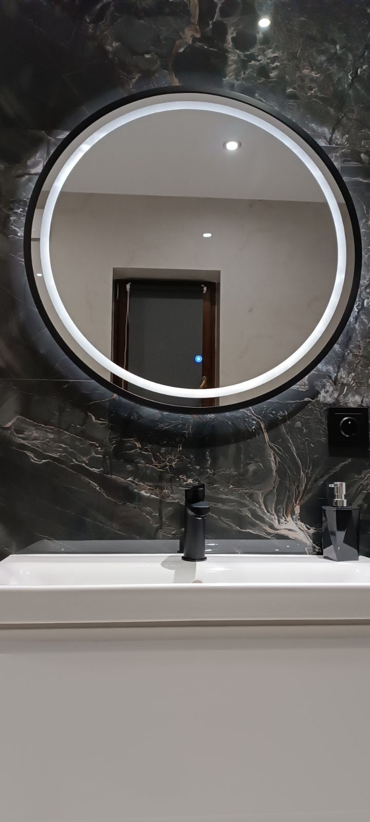 Nowoczesna łazienka z okrągłym lustrem LED, czarną armaturą i marmurową ścianą w odcieniach szarości i czerni.