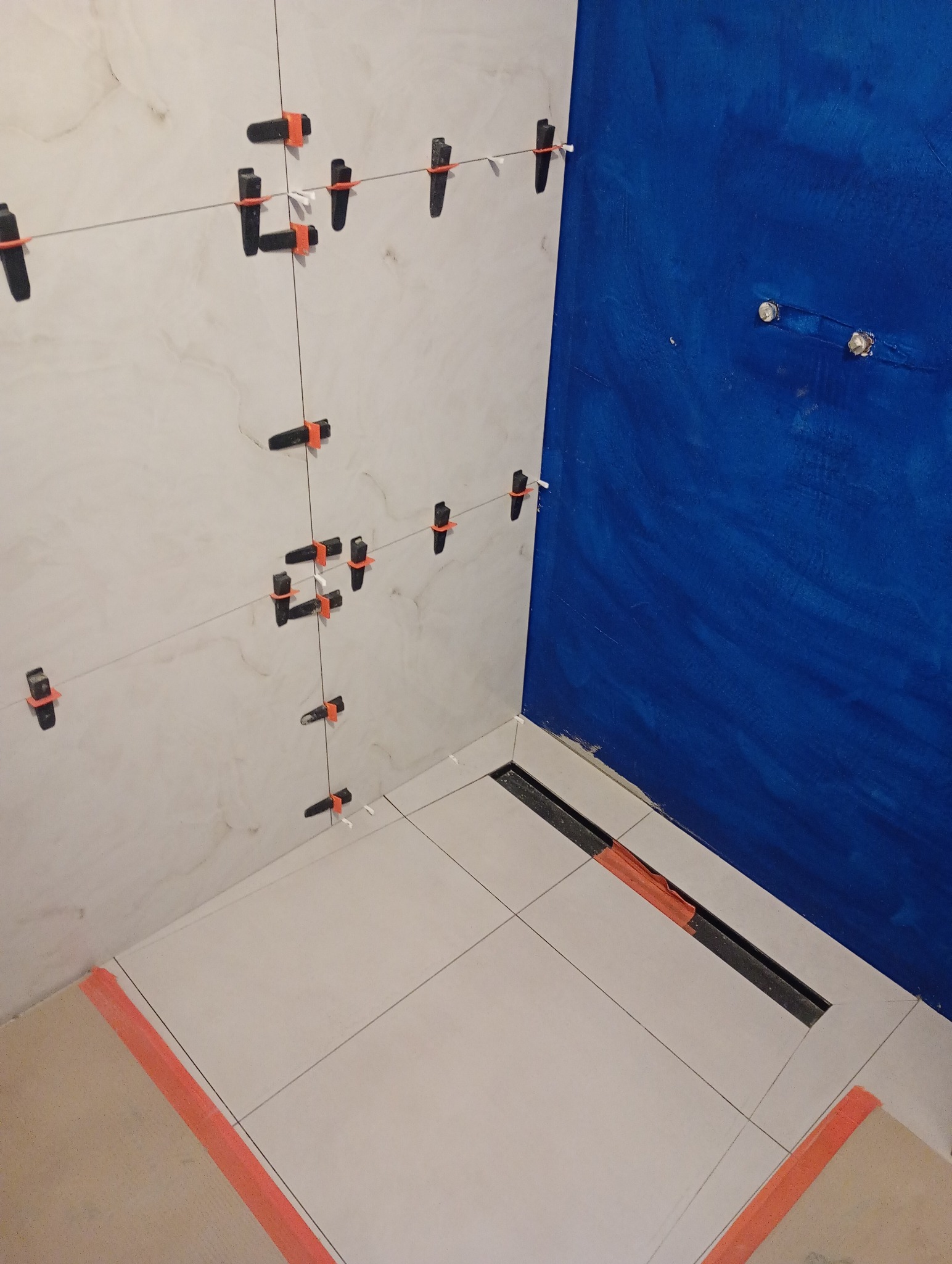 Układanie płytek w rogu łazienki, widoczne klipsy poziomujące i sznurki, świeżo pomalowana na niebiesko ściana obok