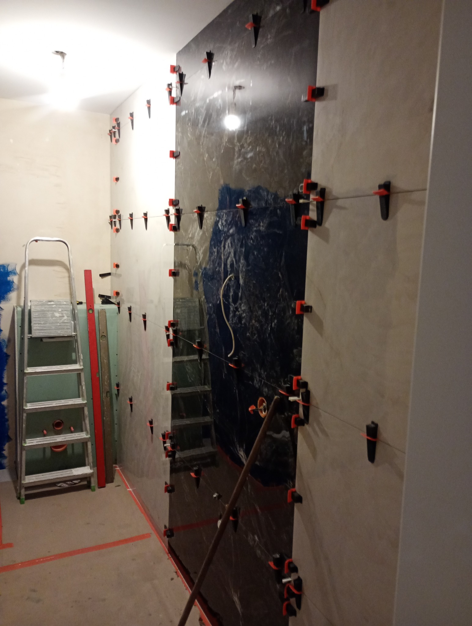 Montaż płytek na ścianie w łazience, widoczne narzędzia glazurnicze, system poziomowania płytek, folia ochronna na części ściany, drabina i poziomica w tle.