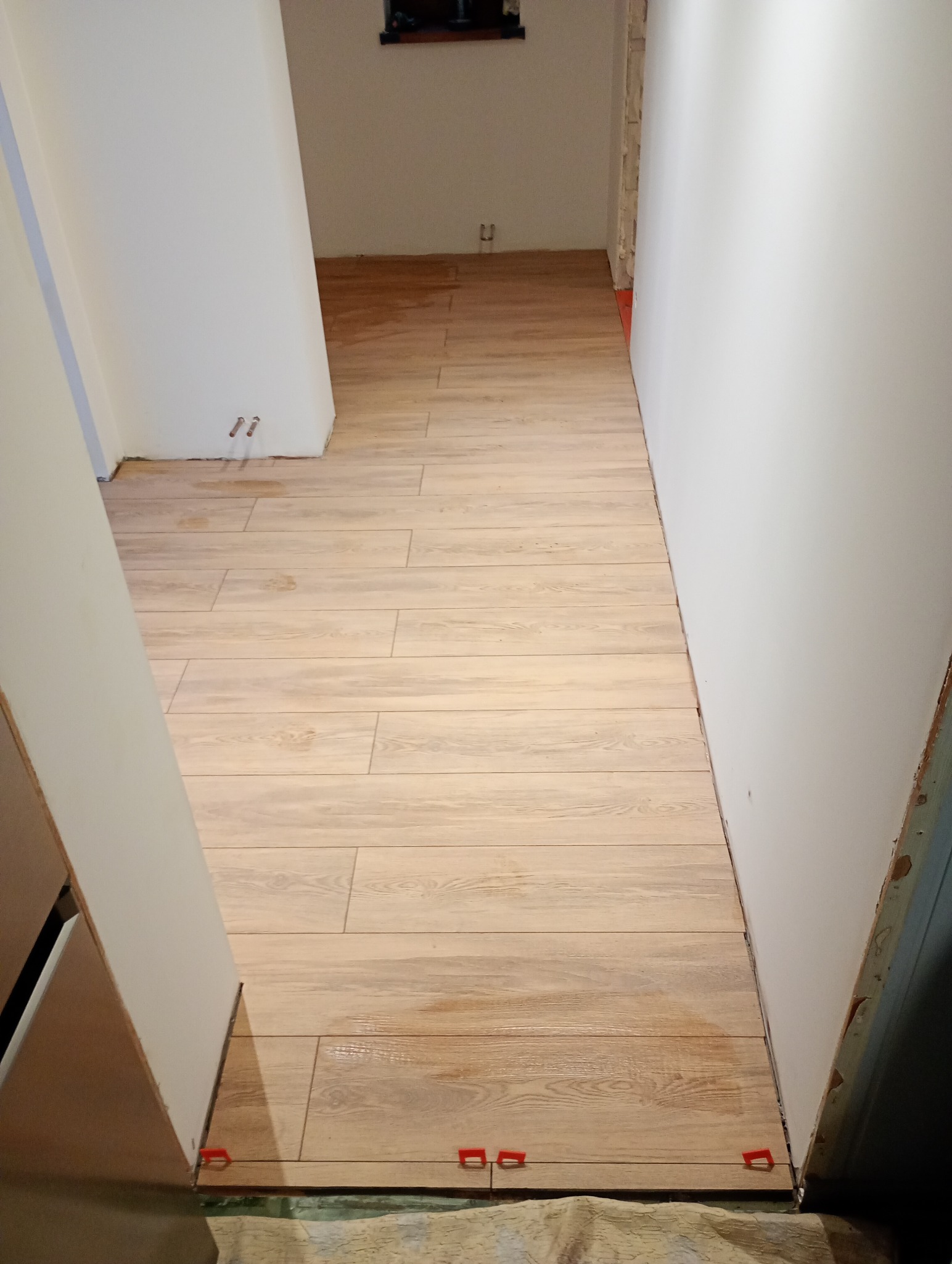 Ułożona podłoga z płytek imitujących drewno w wąskim korytarzu, widoczne czerwone kliny poziomujące, ściany w trakcie malowania.