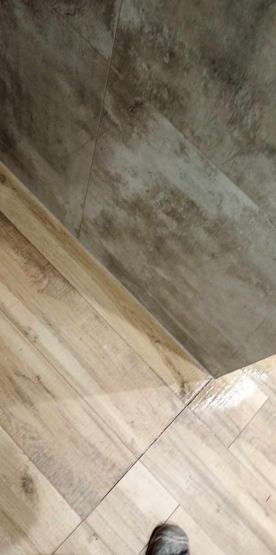 Ułożone płytki podłogowe imitujące drewno, z widocznymi fugami i fragmentem przypodłogowej płytki ściennej w odcieniach szarości, z lekkim połyskiem na powierzchni.